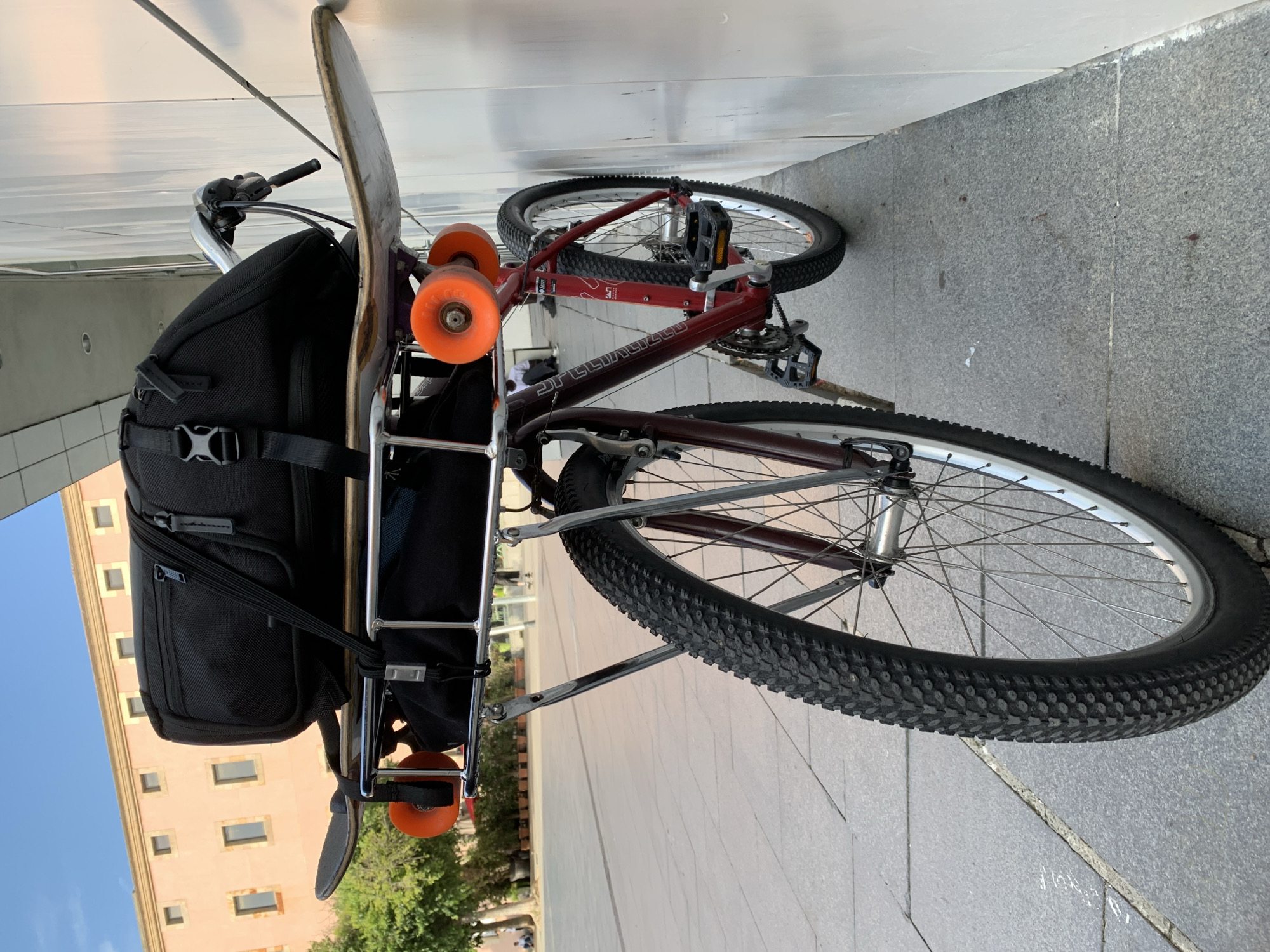 The image shows a Specialized mountain bicycle parked against a light-colored wall. The bike has a maroon frame, silver handlebars, and black knobby tires. A silver rack is mounted over the rear tire. On the rack, there is a black backpack and a skateboard with orange wheels. The bike is on a paved sidewalk with gray tiles. In the background, an office building with windows can be seen under a clear blue sky. It is unlikely that the location is in Barcelona, Spain, due to the architecture of the buildings, which is inconsistent with the typical architecture found in that city. The photograph appears to have been taken during daylight hours.