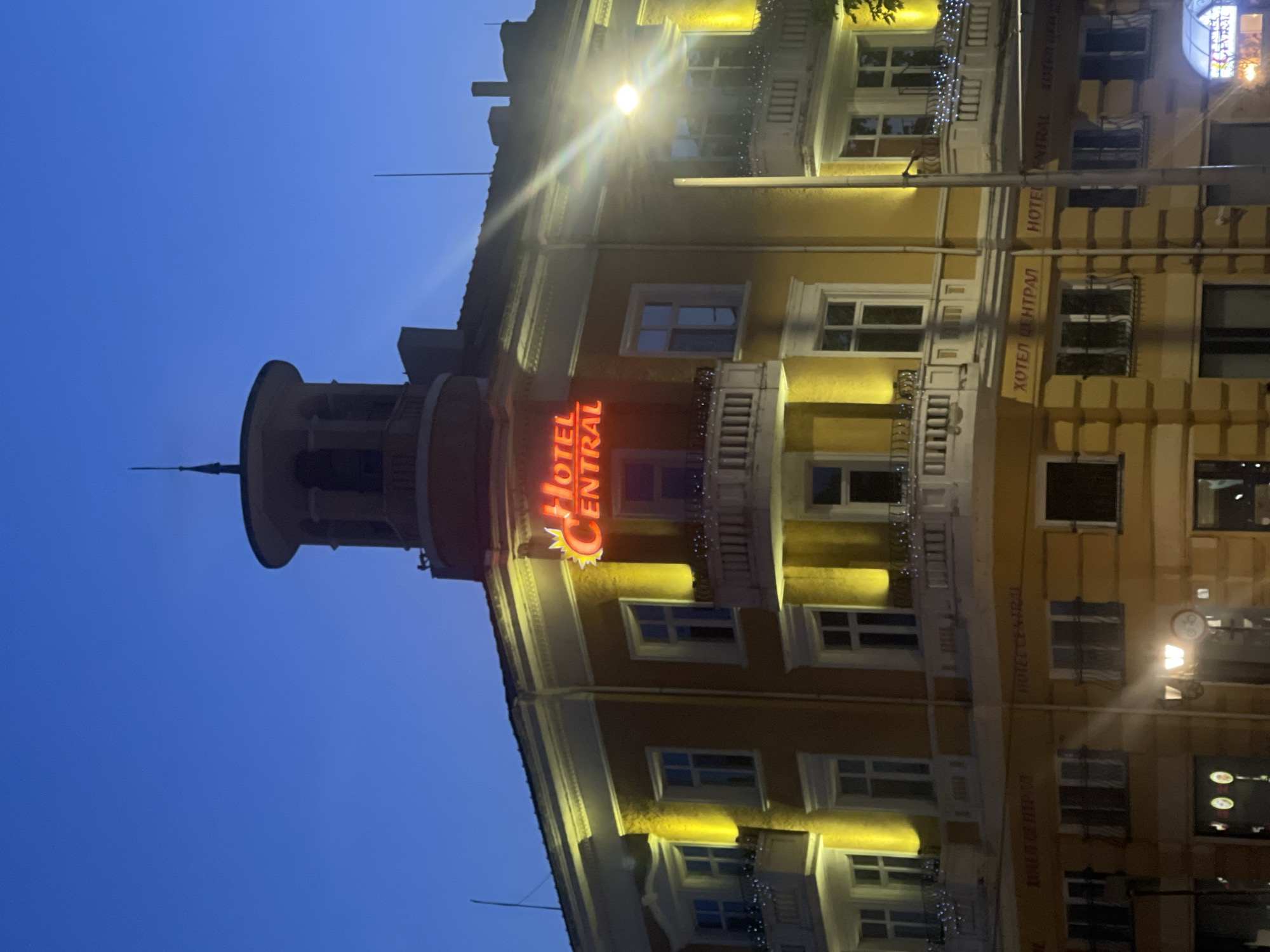 The image shows a building at dusk with a neon sign that reads "HOTEL CENTRAL" in red letters. The building is yellow with white trim and has multiple floors with windows and balconies. The balconies are decorated with fairy lights. There is a tower-like structure on top of the building. The sky is a deep blue. The building is located in Ruse, Bulgaria. In the foreground, there is a street lamp. The scene appears to be captured in an urban environment.