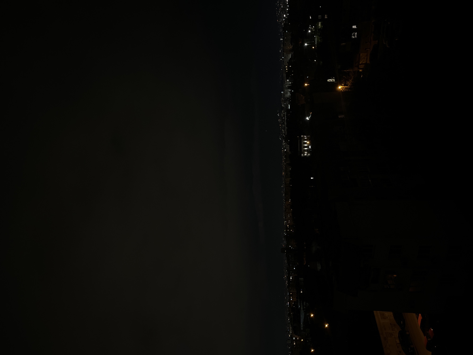 The image shows a nighttime cityscape, possibly Sofia, Bulgaria, viewed from an elevated position. The right side of the image depicts a residential area with lit windows and streetlights. The left side is predominantly dark, suggesting either open space, water, or heavy cloud cover. The lighting is minimal, with illumination primarily from artificial sources within the city. There are few discernible details.