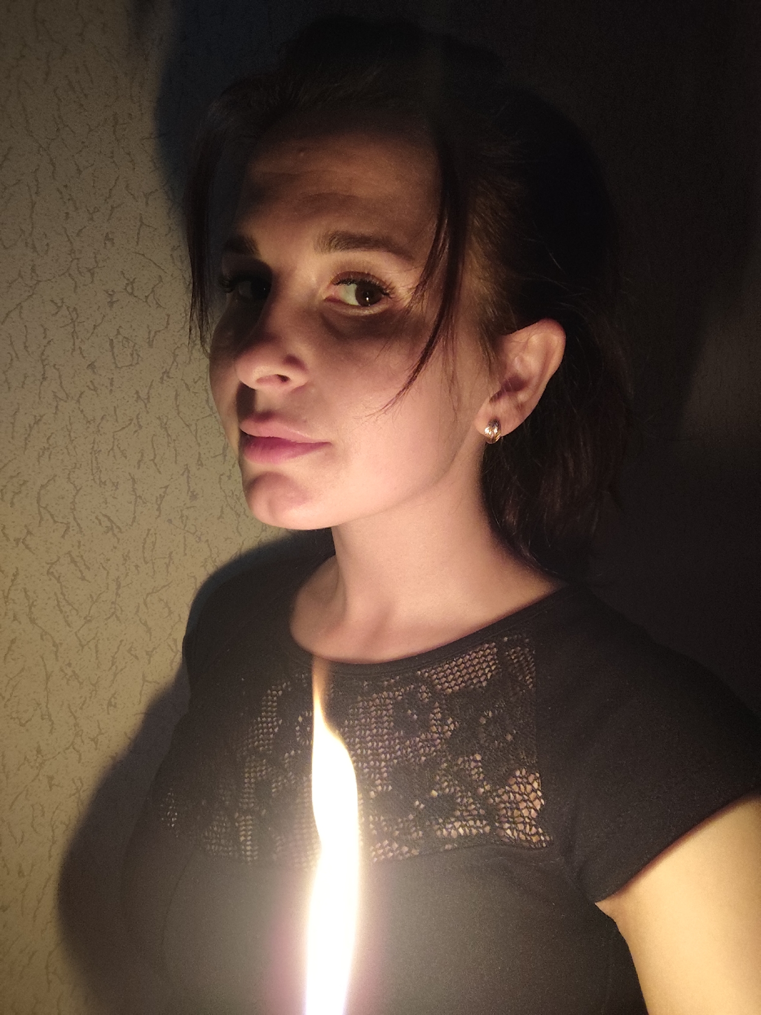 A portrait captures a young woman with dark hair in a dimly lit indoor setting. She is positioned slightly to the right of the frame, gazing directly at the viewer with a calm, somewhat pensive expression. Her features are illuminated primarily by a strong, vertical light source situated low and to her left, which casts dramatic shadows across her face and the background.
The woman has brown eyes, a straight nose, and her lips are slightly parted, showing a hint of pink lipstick. Her dark hair frames her face, with some strands falling forward. She wears a dark top, possibly black, featuring a lace or mesh panel across the upper chest and shoulders. A small, golden hoop earring is visible in her right ear.
The powerful light source, appearing as a bright, glowing streak in the lower center, suggests a candle or similar localized light, creating a high-contrast, moody ambiance. It illuminates the left side of her face and neck, leaving the right in deep shadow.
The background consists of a wall with a textured or subtly patterned wallpaper, likely a light, neutral color, which is partially lit by the same light source on the left, while the right side of the background recedes into darkness. There is no visible text. The overall scene conveys an intimate and somewhat mysterious atmosphere, possibly captured in the evening or night.