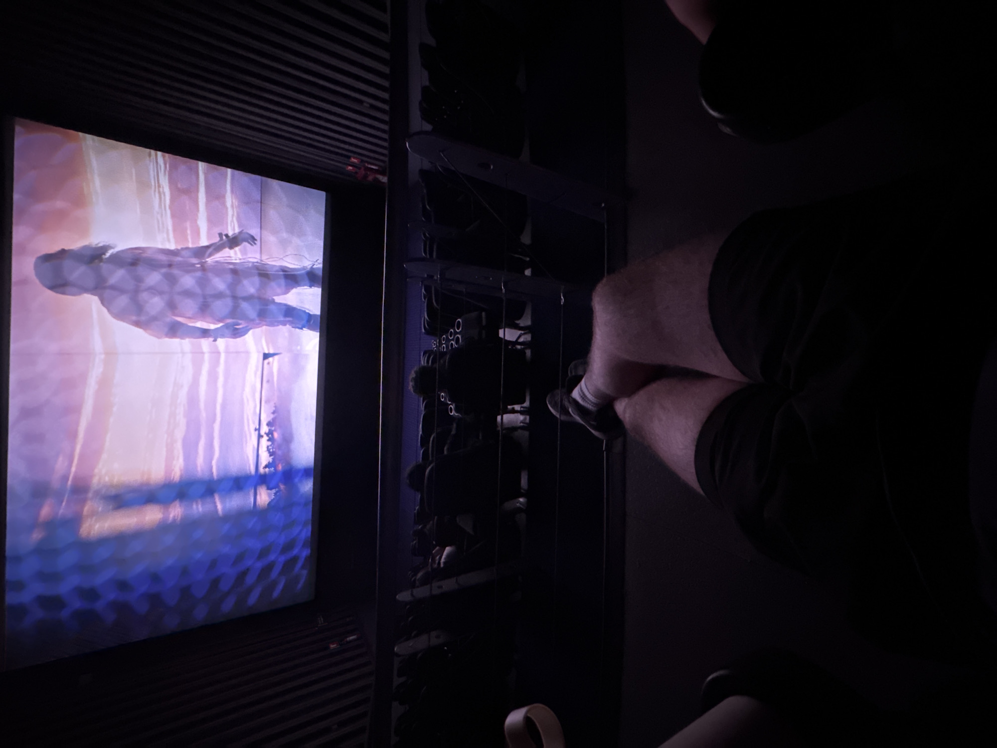 This dimly lit indoor scene captures a person observing a large, vibrant screen displaying digital art. On the right, the lower body of a person is visible, seated with knees bent, wearing dark shorts or trousers and white socks. Their posture suggests they are engrossed in the display.
The prominent feature on the left is a tall, vertical screen showcasing an ethereal, artistic projection. The artwork features a translucent, human-like figure, possibly female, seemingly floating or submerged in water, surrounded by soft shades of lavender, blue, pink, and orange. A subtle, repeating pattern, possibly hexagonal or circular, is superimposed over the figure and the water, adding depth and texture to the digital image.
Between the observer and the screen, a dark, vertical shelving unit or partition stands, its surfaces subtly reflecting the light from the screen. The compartments within this unit are obscure due to the low light, but they appear to hold multiple dark objects, potentially shoes or bags, hinting at a practical storage solution within the space.
The overall setting appears to be an immersive art installation, gallery, or viewing room, designed to focus attention on the glowing screen. The controlled, low lighting enhances the visual impact of the artwork. The scene evokes a quiet, contemplative atmosphere, suggesting a moment of appreciation for the digital art piece. No explicit text is clearly visible in the image. The provided context indicates the location is Puebla, Mexico.