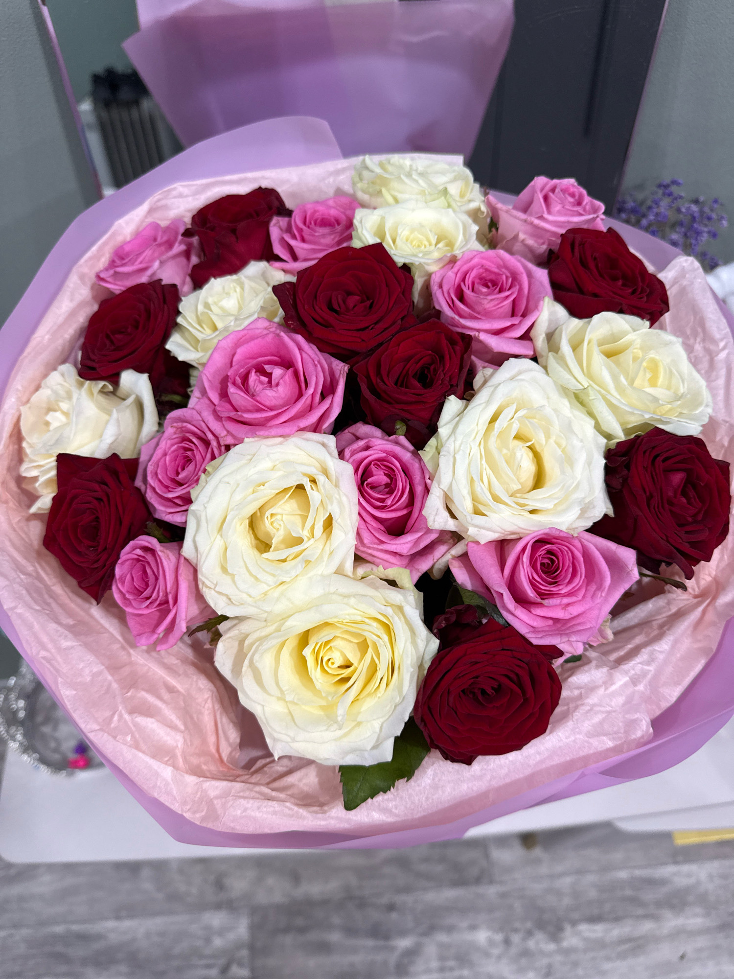 A bouquet of roses is displayed in a pink and purple wrap. The roses are of various colors including red, pink, and cream. The bouquet is placed on a white surface. The background is out of focus but appears to be an indoor setting with muted colors.
