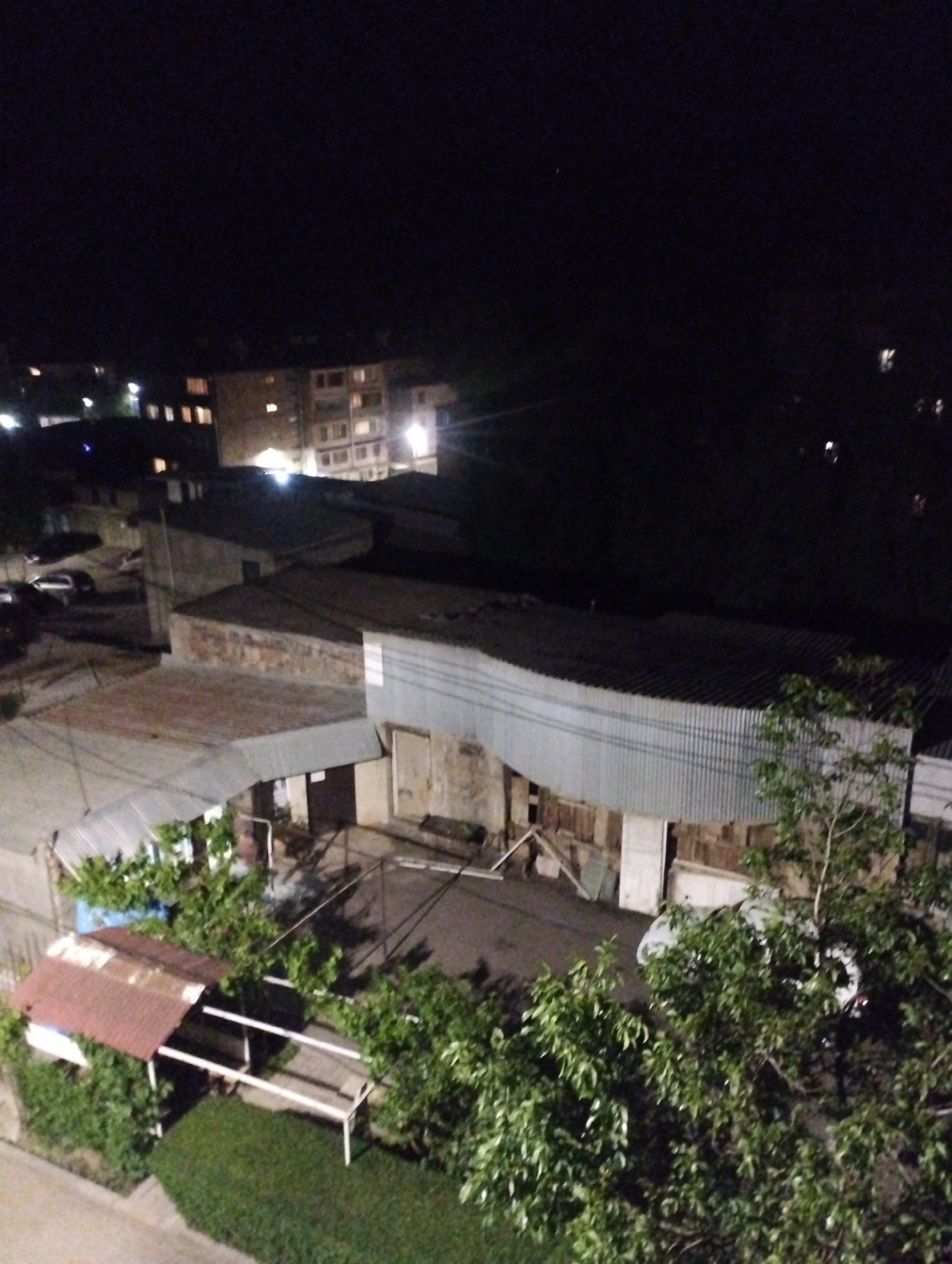 The image provides an elevated nighttime view of a mixed urban and residential area in Yeghegnadzor, Armenia. The scene is characterized by deep darkness punctuated by artificial lights from buildings and possibly streetlights.
In the upper-left and mid-background, a multi-story residential building with several illuminated windows stands out, suggesting habitation. Other smaller buildings with lit windows are also visible further back. The sky above is completely dark, showing no stars or moon, indicating a clear night.
Below the residential building, the landscape consists of various one and two-story structures, many featuring corrugated metal roofs and siding, suggesting a functional or industrial aesthetic for some, or perhaps older construction. These buildings are partially illuminated by nearby lights, casting strong shadows. A light-colored car is parked near some of these structures in the mid-left.
The foreground is dominated by abundant green foliage, including numerous trees and bushes whose leaves are distinct despite the low light. To the lower-left, a small, open-sided shed or shelter with a rusty, reddish-brown corrugated metal roof is visible, positioned next to a neatly kept patch of green grass. Another light-colored car is partially obscured by trees in the lower-right.
There are no visible people or explicit activities taking place, presenting a quiet and still nocturnal scene. The combination of residential and functional structures, along with the natural elements, suggests a lived-in community. No discernible text is visible on any of the buildings or objects.