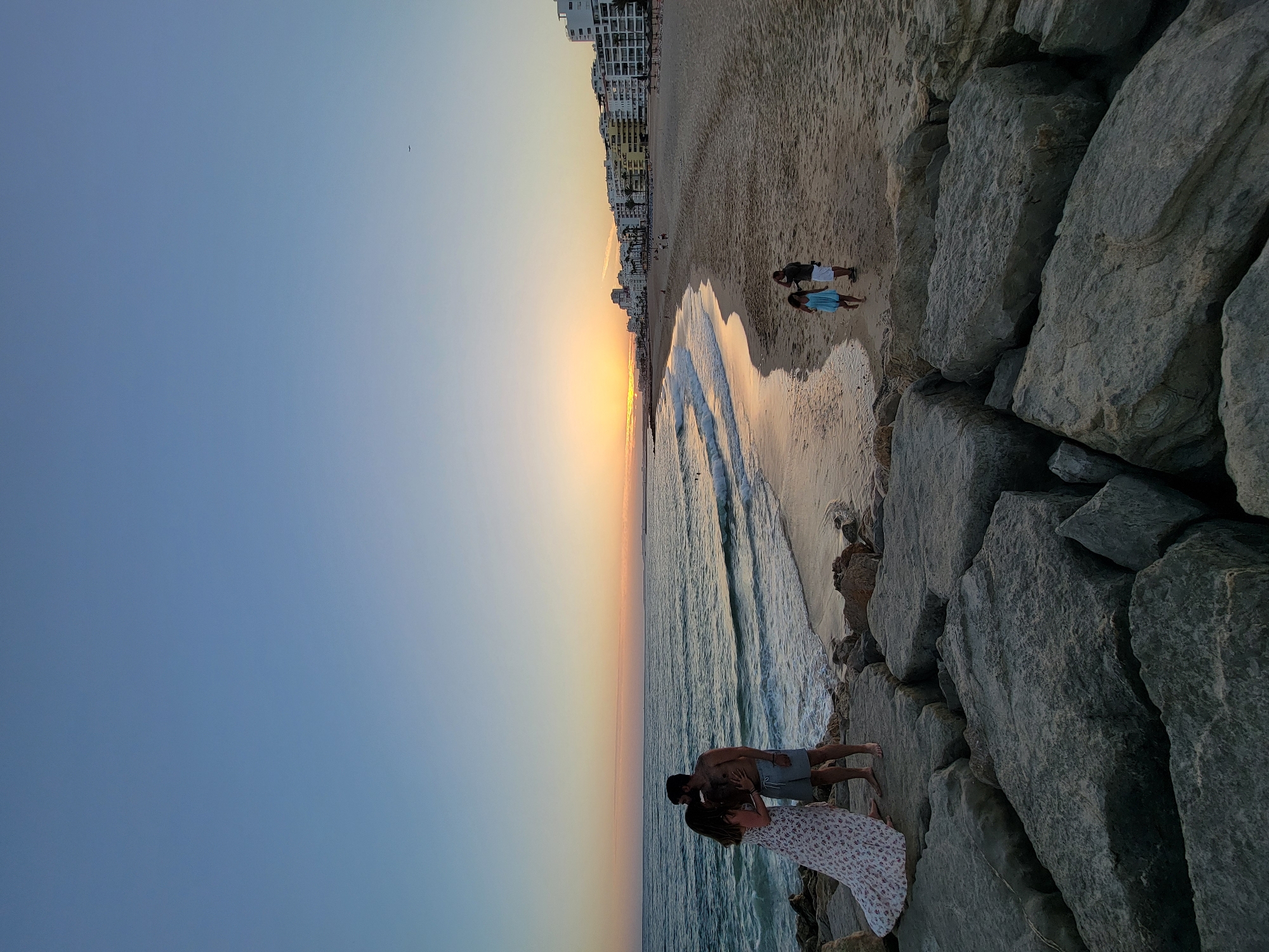 This is a full-frame, vertical, eye-level, outdoor shot of a beach at sunset. In the foreground, large, grey rocks occupy the bottom third of the image. To the left, on the rocks, a couple stands facing each other. The woman, on the left, is wearing a long, white, floral dress and is kissing the man. The man, on the right, is bare-chested and wearing grey shorts. He has his left arm around the woman. The ocean is to the left of the rocks, with waves gently rolling onto the shore. The water is a deep blue, and the wet sand reflects the warm hues of the setting sun. In the mid-ground, a sandy beach stretches horizontally across the image. The sand is light brown, and there are small waves breaking near the shore. Several people are visible on the beach, including a man and a woman walking together and another couple further down the beach. In the background, a line of white buildings, likely hotels or apartments, runs along the coastline, with palm trees interspersed among them. The sky above is a gradient of pale blue at the top, transitioning to soft oranges, pinks, and yellows near the horizon where the sun is setting. A faint airplane is visible in the upper right quadrant of the sky. The scene appears to be set in Quarteira, Portugal, a coastal town known for its beaches and resorts.