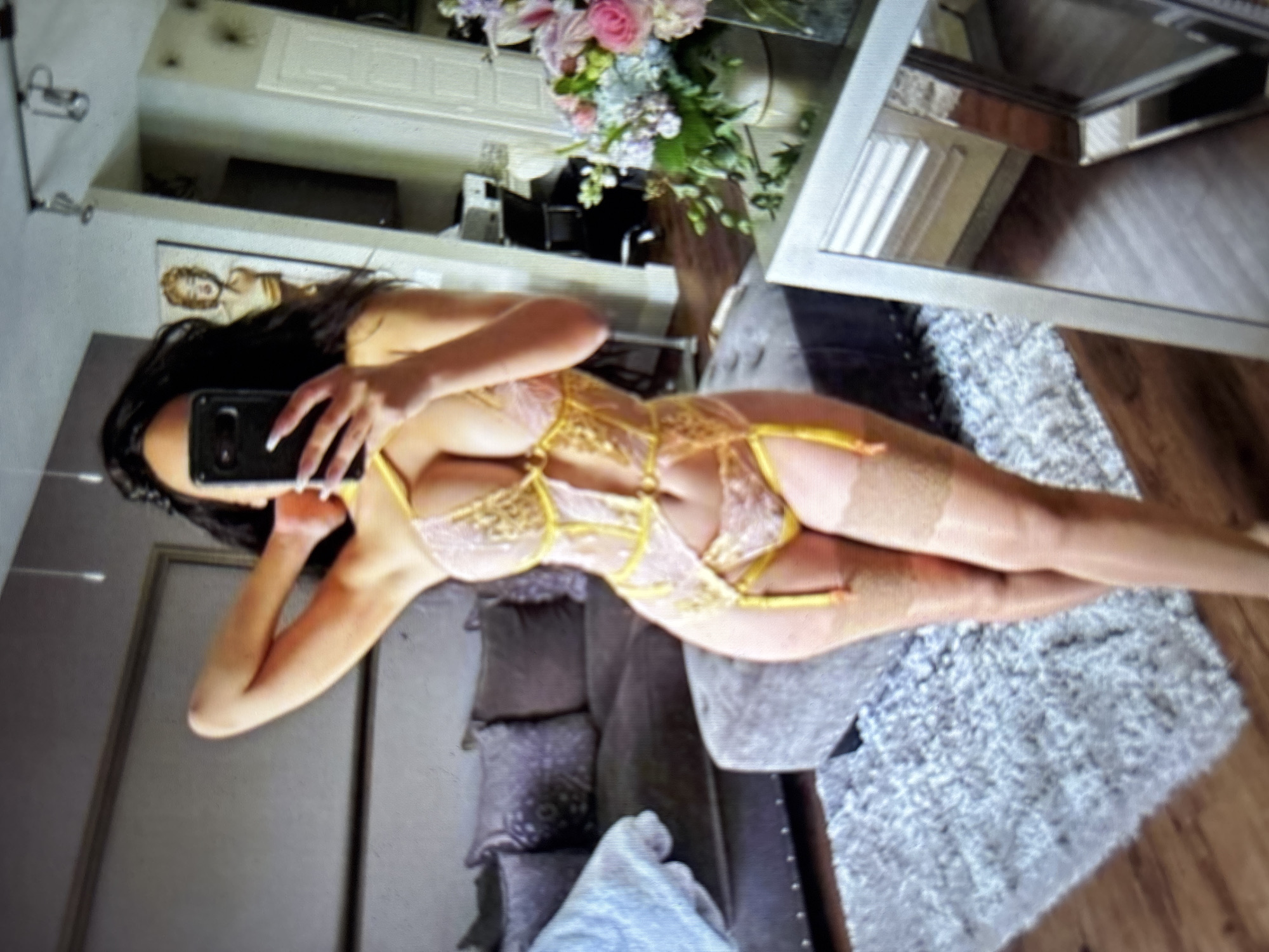 The image shows a woman in lingerie taking a mirror selfie in what appears to be a bedroom. She is wearing a pink and yellow lace bodysuit with matching stockings. The background includes a grey sofa, a fluffy rug, and a bouquet of flowers on a table. The room has wooden floors and a mirror on the wall. The woman is holding a smartphone to take the picture.