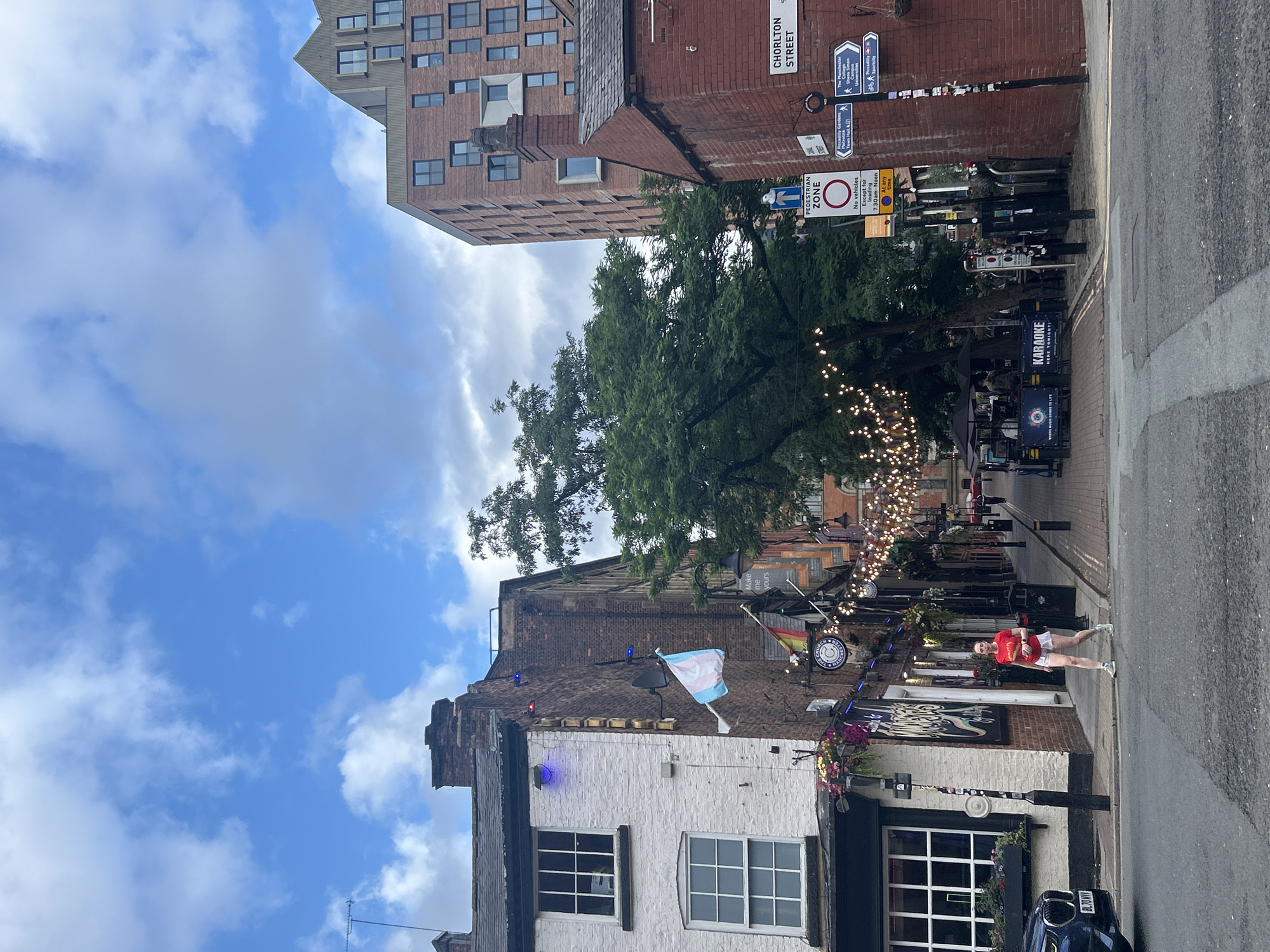 The image shows a street scene in the city of Manchester, United Kingdom. On the left, a white brick building with large windows is visible, with a trans flag hanging from a pole. A person wearing a red t-shirt and white shorts is walking across the street. Further down the street, string lights are hung across the road, illuminating an outdoor seating area with umbrellas and tables. A sign for "Karaoke" is visible. On the right, a brick building displays a street sign that reads "Chorlton Street." A pedestrian zone sign is also present. In the background, modern buildings rise above the older structures. The sky is blue with scattered clouds.