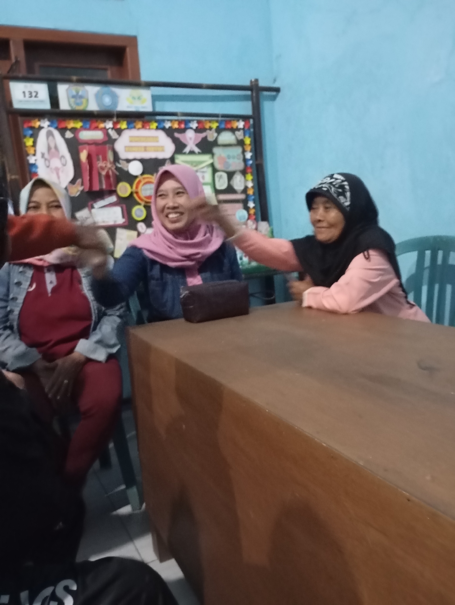 The image depicts an indoor scene in Ketepung, Indonesia, likely a community gathering or a classroom. Three women are the main subjects. On the left, a woman wearing a light blue denim jacket over a maroon top and red pants is visible from the chest down, her arms extended forward. In the center, a woman in a pink hijab and a blue denim jacket is smiling broadly, also with her arms extended. To the right, an older woman in a black hijab and a pink shirt is laughing, her right arm raised. They appear to be interacting, possibly high-fiving or playing a game. A brown wooden table dominates the foreground. In the background, a bulletin board adorned with colorful decorations, cutouts of people and ribbons, and text is mounted on a light blue wall. A metal rack supports the bulletin board. A chair is partially visible behind the older woman. The lighting is dim, suggesting an evening setting.