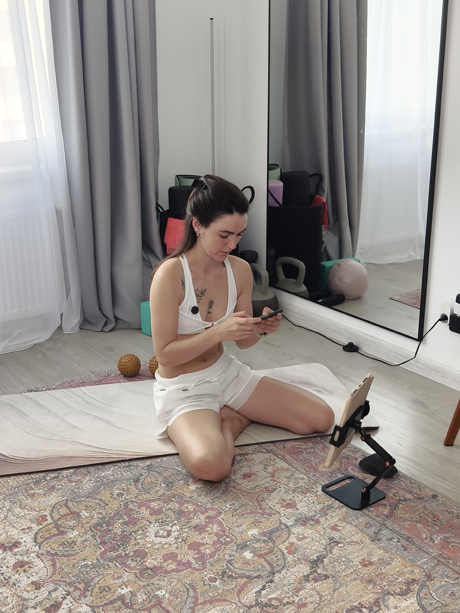 In a room with light-colored walls and wooden flooring, a woman in a white sports bra and white shorts is seated on a yoga mat. The mat is placed on top of a patterned rug. She is holding a smartphone in her hands, looking down at it. To her right, on the mat, is a tablet mounted on a black stand. In the background, to the left, there are grey curtains. To the right, a large mirror reflects the room, showing various fitness equipment, including kettlebells and a pink exercise ball, and more grey curtains. The room appears to be a personal exercise space. There is no specific information to indicate the location is in Tyumen, Russia.