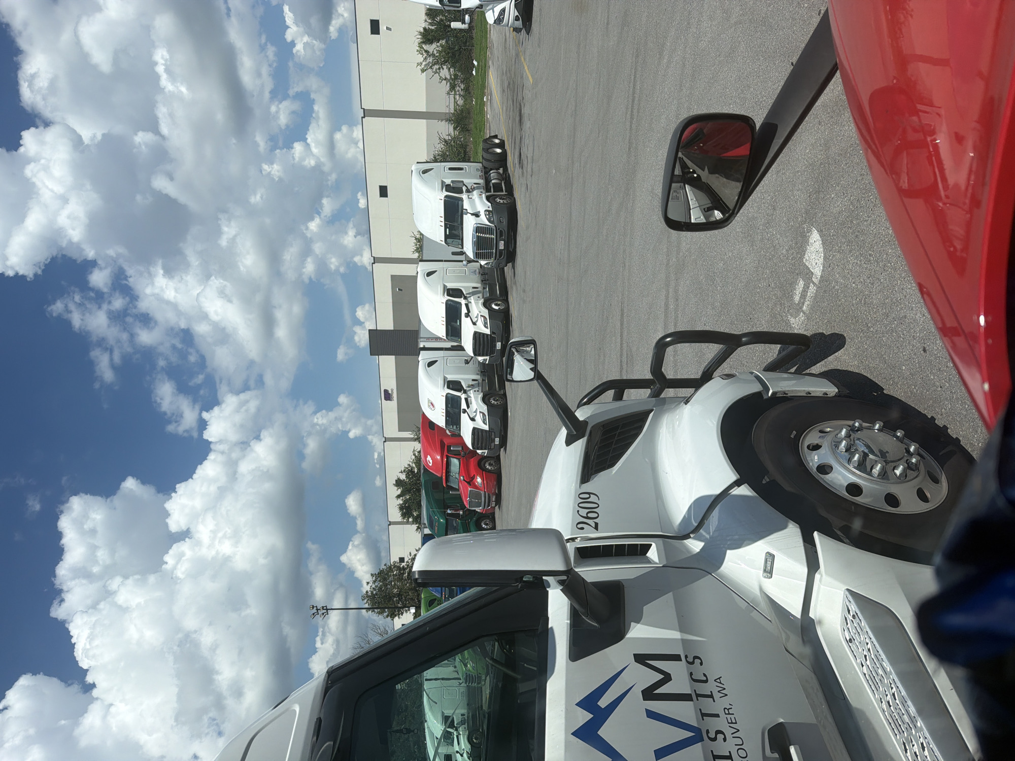 A fleet of white and red semi-trucks are parked in a lot with a warehouse in the background under a partly cloudy sky. The trucks are arranged in rows, with some on the left side of the image in the foreground and middle ground, and others in the middle and right ground. The truck in the immediate foreground on the left is white with blue and black lettering that says "VM LOGISTICS VANCOUVER, WA". A red semi-truck is parked to the right of the foreground truck, and further back, several more white and red semi-trucks are visible. The warehouse in the background is a large, beige structure with dark trim and windows. The ground is paved asphalt, and there are some trees and bushes visible in the distance. A traffic light is visible in the background.