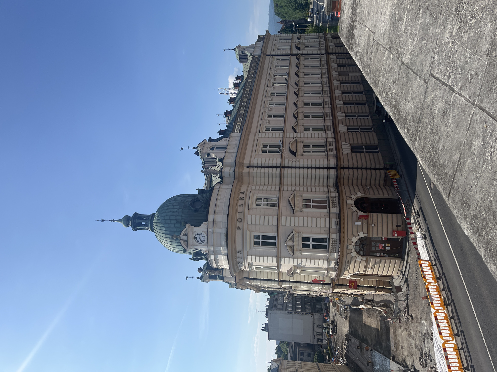 A wide shot captures a prominent, historic building with a green dome and a clock on its facade, standing on a corner in an urban environment. The building features ornate architectural details, including columns, arched doorways, and numerous windows. A sign on the building reads "POCZTA POLSKA." In front of the building, a street is visible, with construction barriers and some ongoing roadwork. Adjacent to the building, other structures and a clear blue sky are present. The image is taken from a slightly elevated perspective, looking down on the street and foreground.