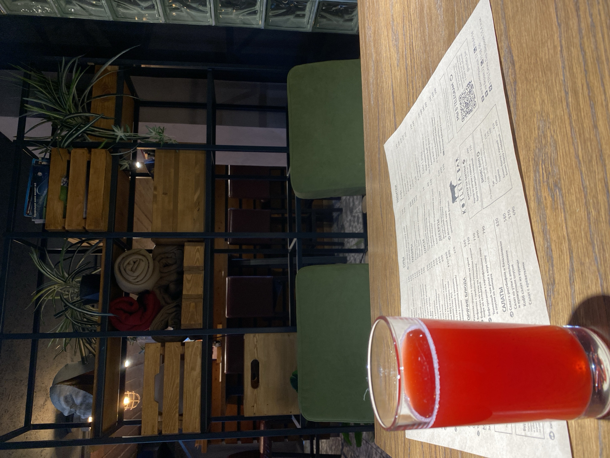 The image depicts a restaurant setting. A glass of red liquid, possibly juice or a cocktail, sits on a wooden table alongside a menu. The menu is from a restaurant called "Kotleta Bar", with locations in Izhevsk, Russia, specifically on Krasnogeroyskaya Street, Bersha Street, and in the Sigma Shopping Center. The menu lists various dishes, including burgers, salads, and soups. In the background, there's a structural element made of dark metal framing, interspersed with wooden accents. There are green upholstered chairs and decorative plants arranged in wooden crates. The overall impression is of a modern, casually stylish eatery.