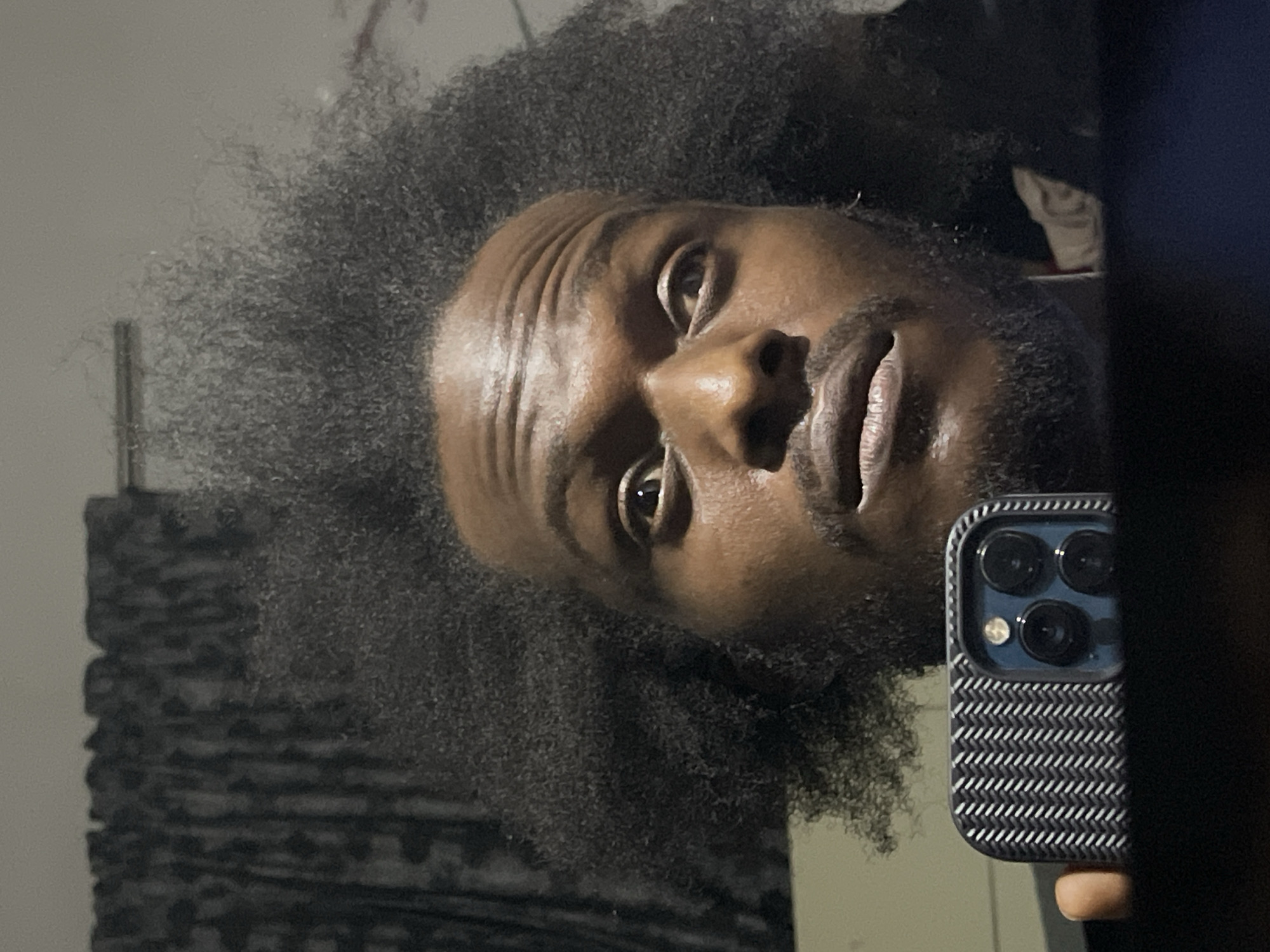 A close-up selfie of a Black male with dark skin and full, natural hair. He has a short beard and mustache. His eyes are open and appear to be looking at the camera. He is holding a smartphone with a patterned case, partially obscuring the right side of his face and the scene. The background is blurred, suggesting an indoor setting with neutral-colored walls. The orientation of the image is rotated. There is no indication in the image that reveals a specific location in Kuchingoro, Nigeria.