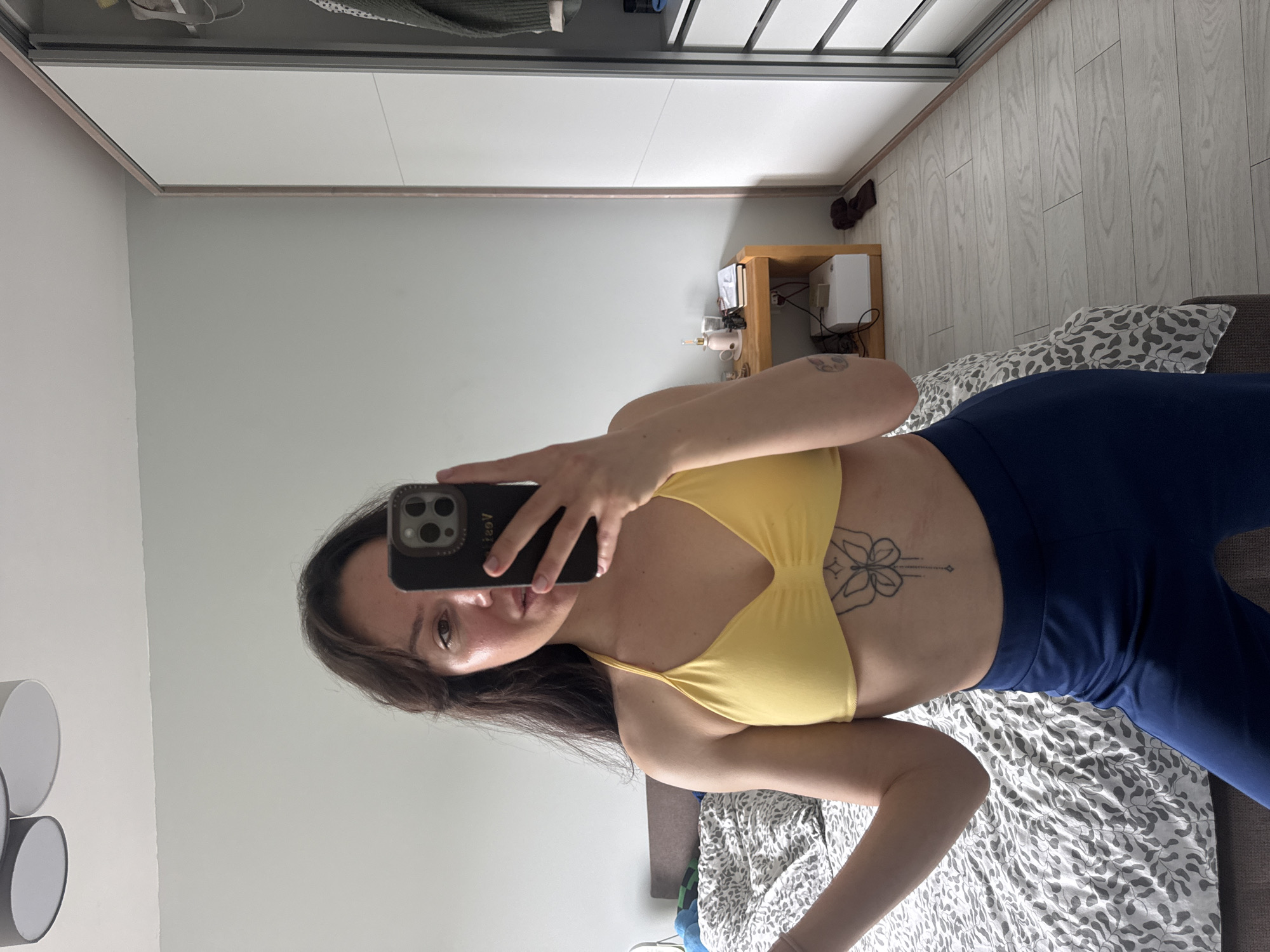 A woman is taking a selfie in a room with a bed, wardrobe, and bedside table. She is wearing a yellow sports bra and blue leggings. The room appears to be indoors. The location is not discernible from the image.