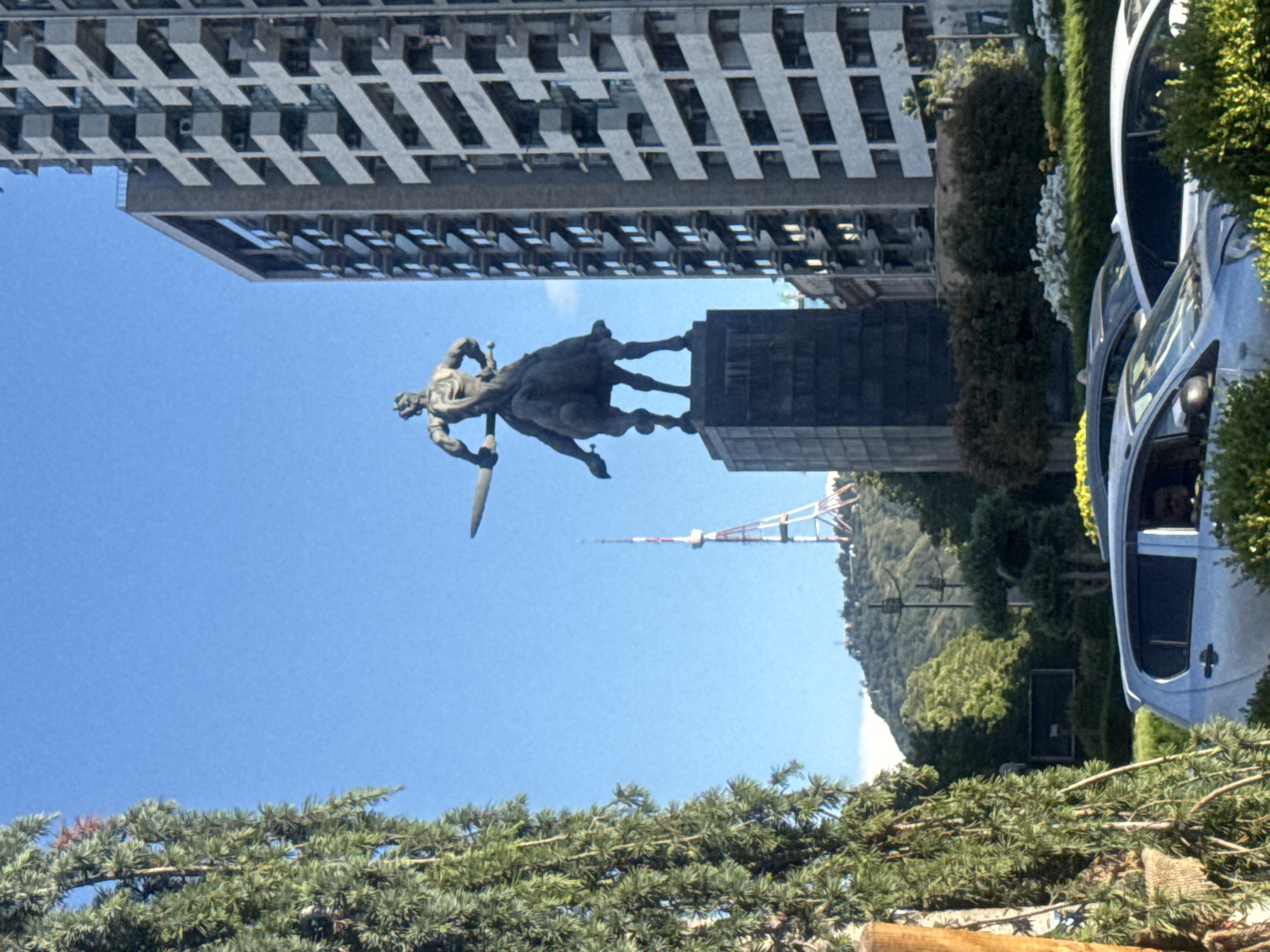 This is a medium-shot, eye-level, outdoor photograph taken on a clear, sunny day in Tbilisi, Georgia, featuring a statue of a warrior on horseback in the foreground, a tall modern building on the right, and a television tower in the background.
The statue, made of dark metal, depicts a muscular male figure wearing a helmet and holding a sword raised in his right hand, mounted on a powerful horse. The statue stands on a tall, dark, rectangular pedestal.
To the right of the statue, a multi-story building with a grey, concrete facade and numerous balconies dominates the right side of the frame. The building appears to be residential or an office complex.
In the mid-ground, a tall, red and white lattice television tower is visible on a distant hill. The hill is covered with lush green trees and vegetation.
In the lower foreground, the top of a light blue sedan is visible, along with other cars and greenery. A large, dense green tree with needle-like leaves is prominently positioned on the left side of the frame, partially obscuring the view of the background. The sky is a clear, bright blue. The overall scene suggests an urban environment with significant landmarks.
