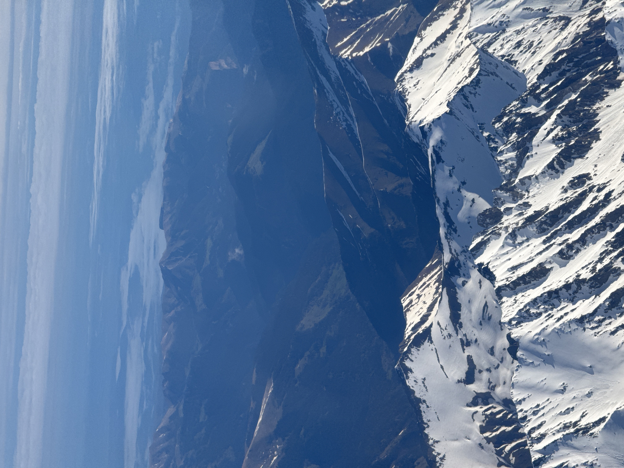 The image depicts a mountainous landscape with significant elevation changes. The left side of the image shows a valley filled with a hazy blue atmosphere, possibly due to atmospheric perspective or low-lying clouds. Adjacent to the hazy valley is a series of dark, steep mountain slopes with hints of green vegetation. The right side of the image features mountains covered in snow, with alternating bands of snow and bare rock or exposed soil due to the terrain's ridges and valleys. The overall impression is of a rugged, high-altitude environment. Given the reference to Sochi, Russia, it is likely that this image depicts the Caucasus Mountains, known for their steep slopes and snowy peaks.