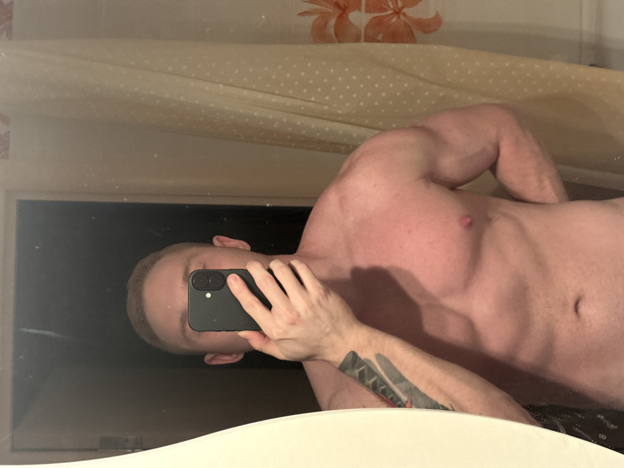 A shirtless man is taking a selfie in a bathroom mirror. The bathroom has a shower curtain with an orange floral pattern and a beige polka dot pattern. The man has a tattoo on his left forearm. The location is likely in Chaikovsky, Russia. The man is holding a black smartphone. His face is partially obscured by the phone.
