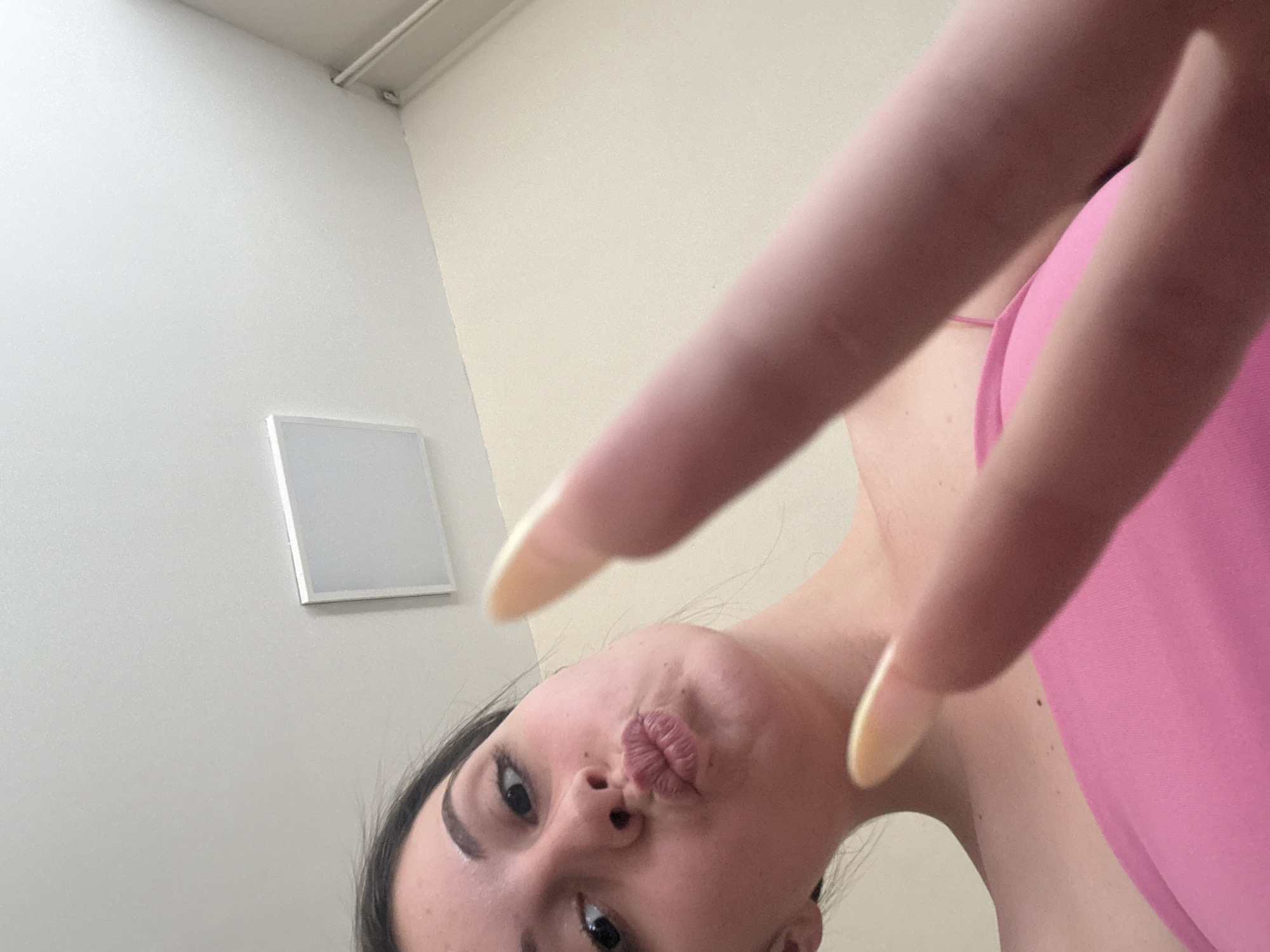 A low-angle, close-up shot captures a young woman with dark hair, making a kissy face. She is wearing a pink top and her right hand is raised, with two fingers extended in a peace sign. Her long, light-colored fingernails are visible. The background consists of a plain, light-colored wall and ceiling with a square light fixture. No specific location in Burgas, Bulgaria, can be identified from the image.