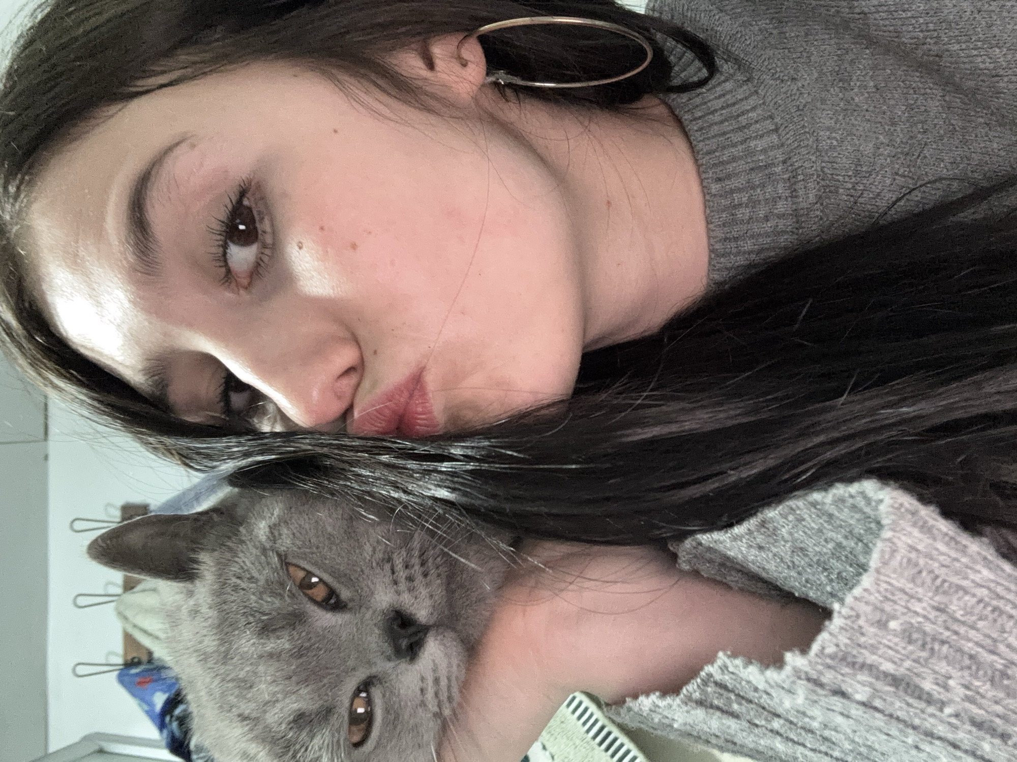 An intimate, close-up indoor portrait features a young woman and a grey British Shorthair cat, likely taken as a selfie from an overhead or angled perspective. The woman, with long dark hair, occupies the upper and central part of the frame, lying on her side. She is wearing a grey knitted sweater and has a large, thin hoop earring visible on her right ear. Her face shows natural makeup, with visible eyelashes and some freckles. She is making a slight pouting or kissing face towards the camera, suggesting affection.
Cradled beside her, partially obscured by her hair and body, is a grey cat with amber-colored eyes, characteristic of the British Shorthair breed. The cat's head and part of its body are visible, and it appears calm, looking directly towards the camera. The woman's arm or hand is visible, gently holding the cat.
The background is softly blurred but indicates an indoor setting, possibly a home, with hints of light-colored walls and what might be hangers or hooks in the upper left corner. The overall mood of the image conveys warmth and companionship between the woman and her pet. No visible text is present in the image. This scene takes place in Фоки, Russia.