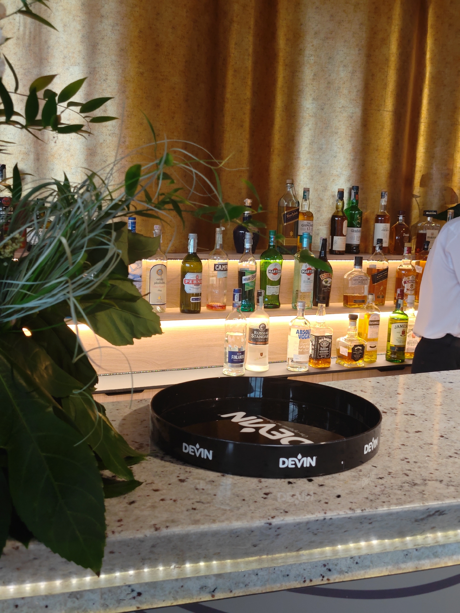 The image shows a bar counter with various liquor bottles on a shelf behind it. Brands like Johnnie Walker Blue Label, Glenlivet, Glenfiddich, Chivas Regal, Jack Daniel's, and Absolut Vodka are visible. A black tray with "Devin" written on it sits on the countertop. Part of a plant is in the foreground. A person wearing a white shirt is partially visible on the right. The image's environment looks to be indoors, possibly in a bar or restaurant. Devin is a Bulgarian mineral water brand. Given that and the Cyrillic characters on the tray, the location is likely in Bulgaria, possibly in the city of Varna.