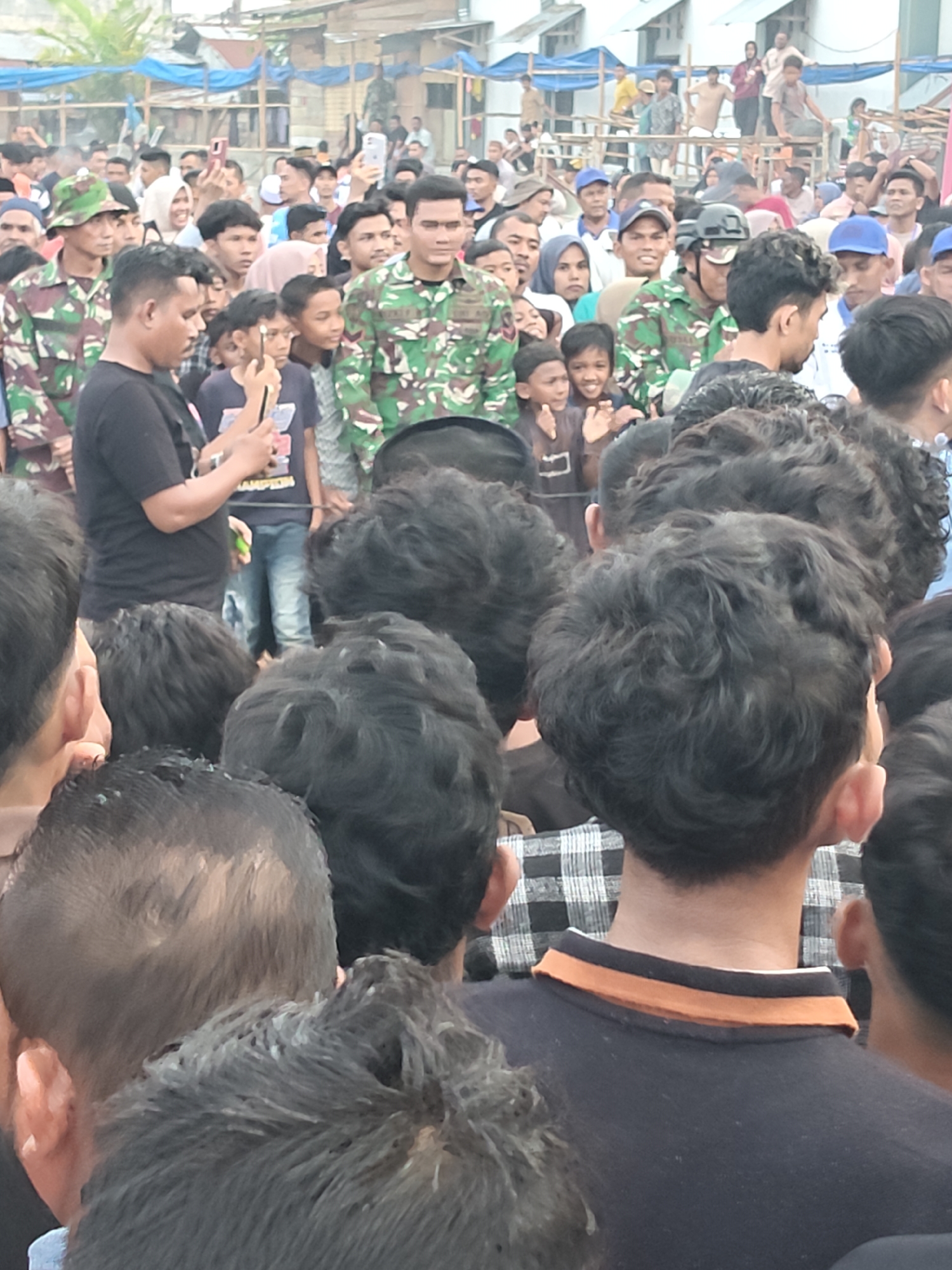 The image captures a lively outdoor gathering in Desa Meunasah Blang, Indonesia. A crowd of people, predominantly young men and boys, are densely packed together, suggesting a significant event or popular attraction. Many are looking in the same direction, with some holding up smartphones, possibly to capture the moment or a performance.
In the center, a man in a camouflage uniform, likely a soldier, stands out. He is the focal point for some of the onlookers. The atmosphere appears energetic and engaged. The time of day seems to be daytime, with natural light illuminating the scene. The background reveals a mix of structures, including what appear to be temporary shelters or stalls, perhaps indicating a local market, festival, or community event. The presence of blue tarps suggests some form of temporary roofing or shading.
The clothing of the individuals is varied, from casual t-shirts to the military uniform. The hairstyles are diverse, with many young men sporting curly or short cuts. The overall impression is one of a busy public assembly, possibly a cultural celebration, a gathering of community members, or a military-related event drawing local attention. No specific text is clearly legible in the image.