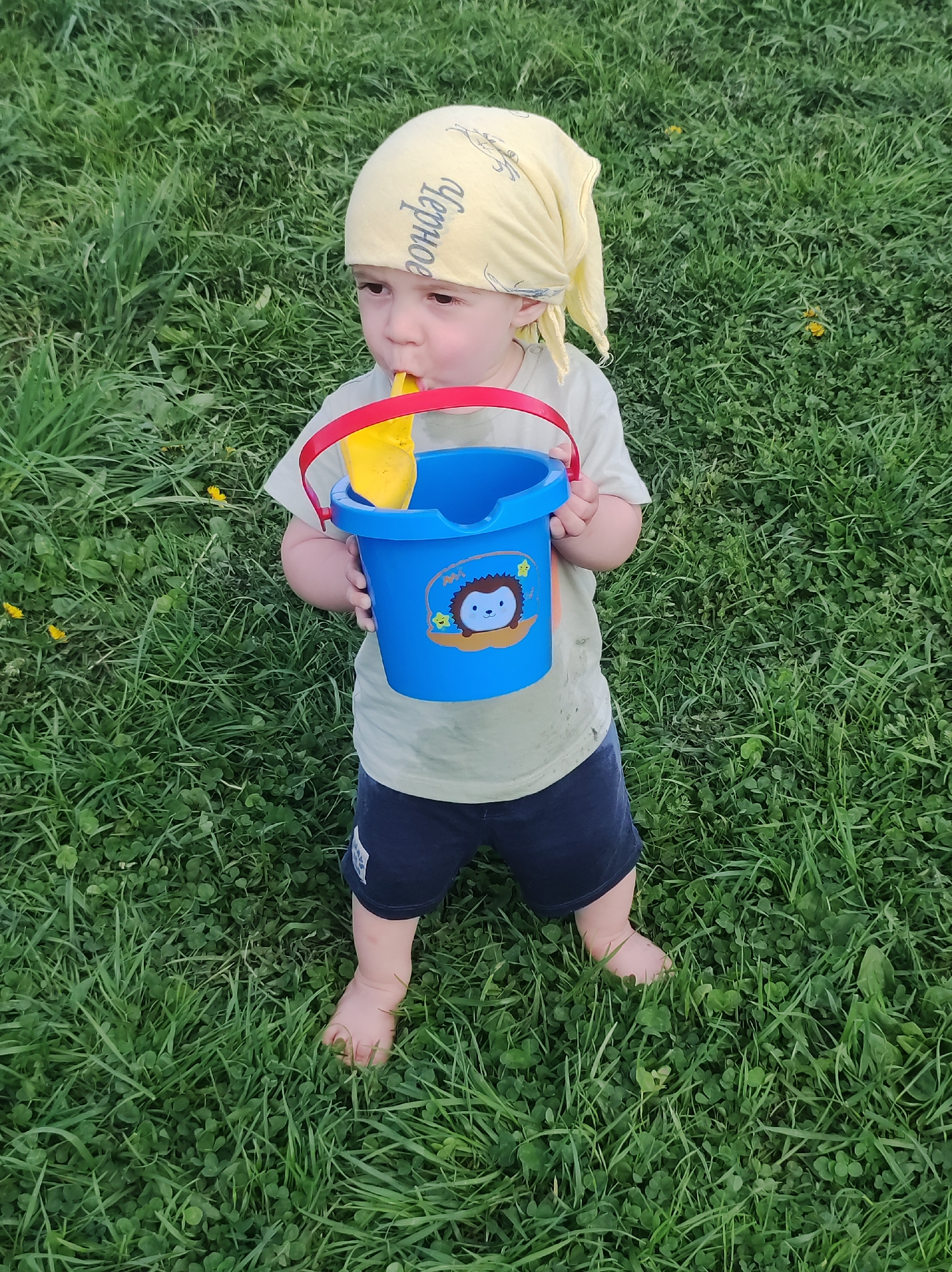 A young child stands barefoot on a patch of green grass. The child is holding a blue plastic bucket with a red handle; a yellow plastic shovel is inside the bucket. The child has a yellow bandana on their head. The exact location in the city of Фоки, Russia, cannot be determined from the visual information alone.