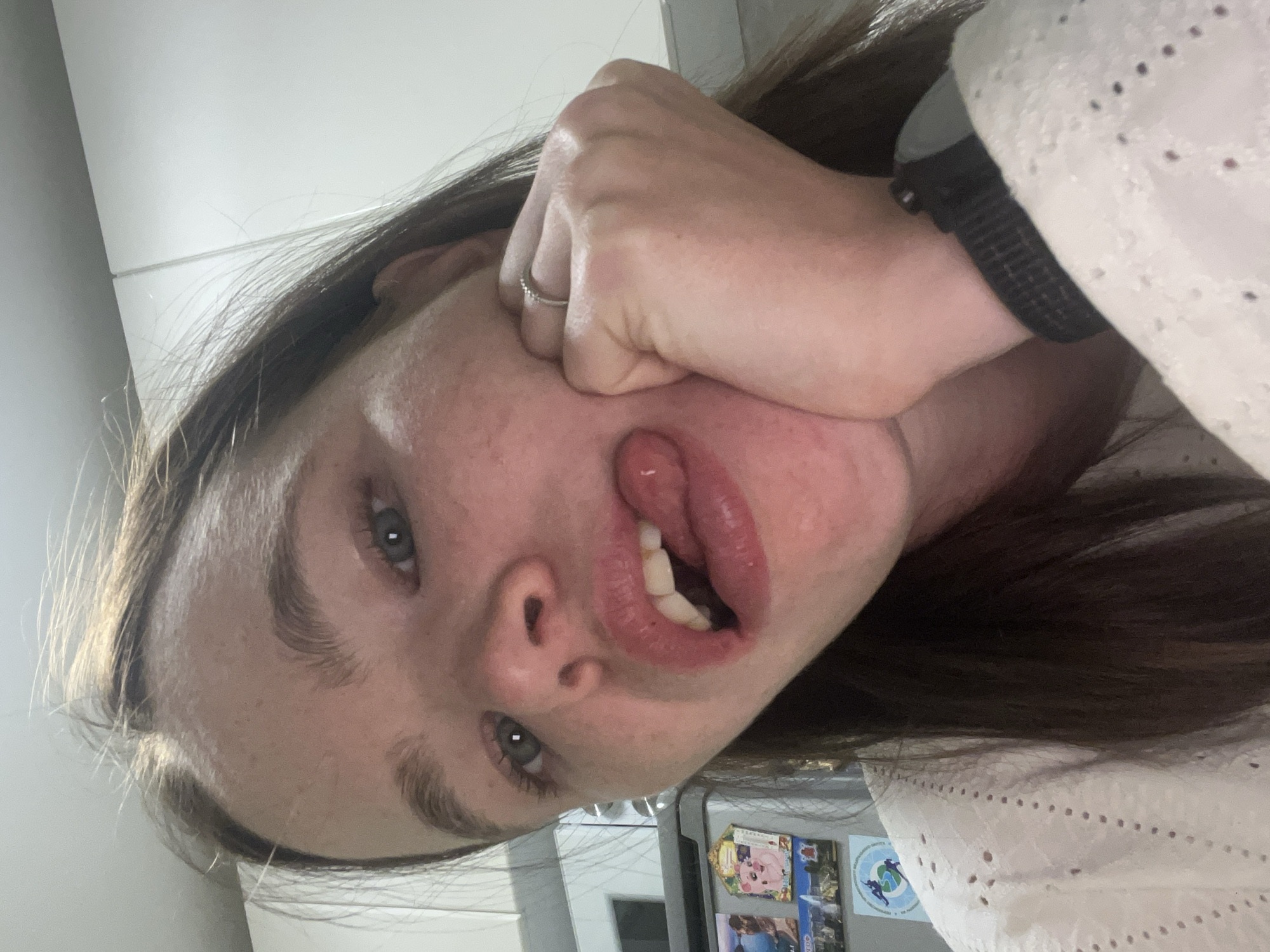 A woman is posing for a close-up, low-angle, selfie. She is holding her cheek with her hand, distorting her face. She has brown hair and blue eyes. She is wearing a white, lacy blouse and a watch with a black band. A silver ring is visible on her left ring finger. Behind her is a white refrigerator with various magnets attached. Based on information from the magnets visible on the refrigerator, the location may be Chaikovsky, Russia.