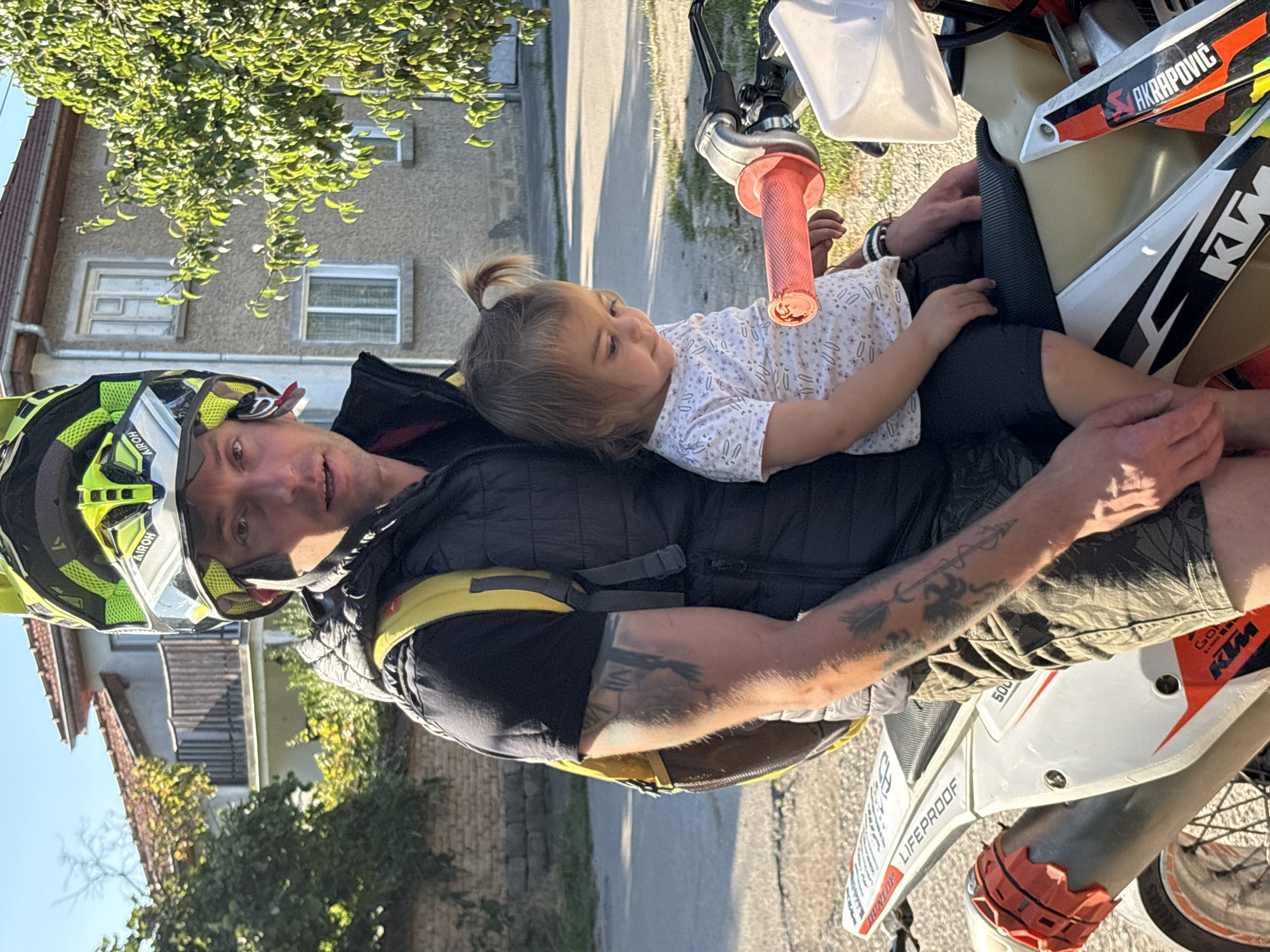 In Oreshak, Bulgaria, a man wearing a yellow and black motocross helmet and a black puffer vest over a black t-shirt is seated on a white and orange dirt bike. He has a yellow backpack on. A young child with a ponytail is sitting in front of him on the dirt bike, wearing a white t-shirt with a repeating pattern of small, light-colored objects. The child is looking down. The man has tattoos on his left arm, including a roman numeral and an arrow design. The dirt bike has "LIFEPROOF" and "KTM" branding. The background shows a house with a balcony and trees, illuminated by sunlight.