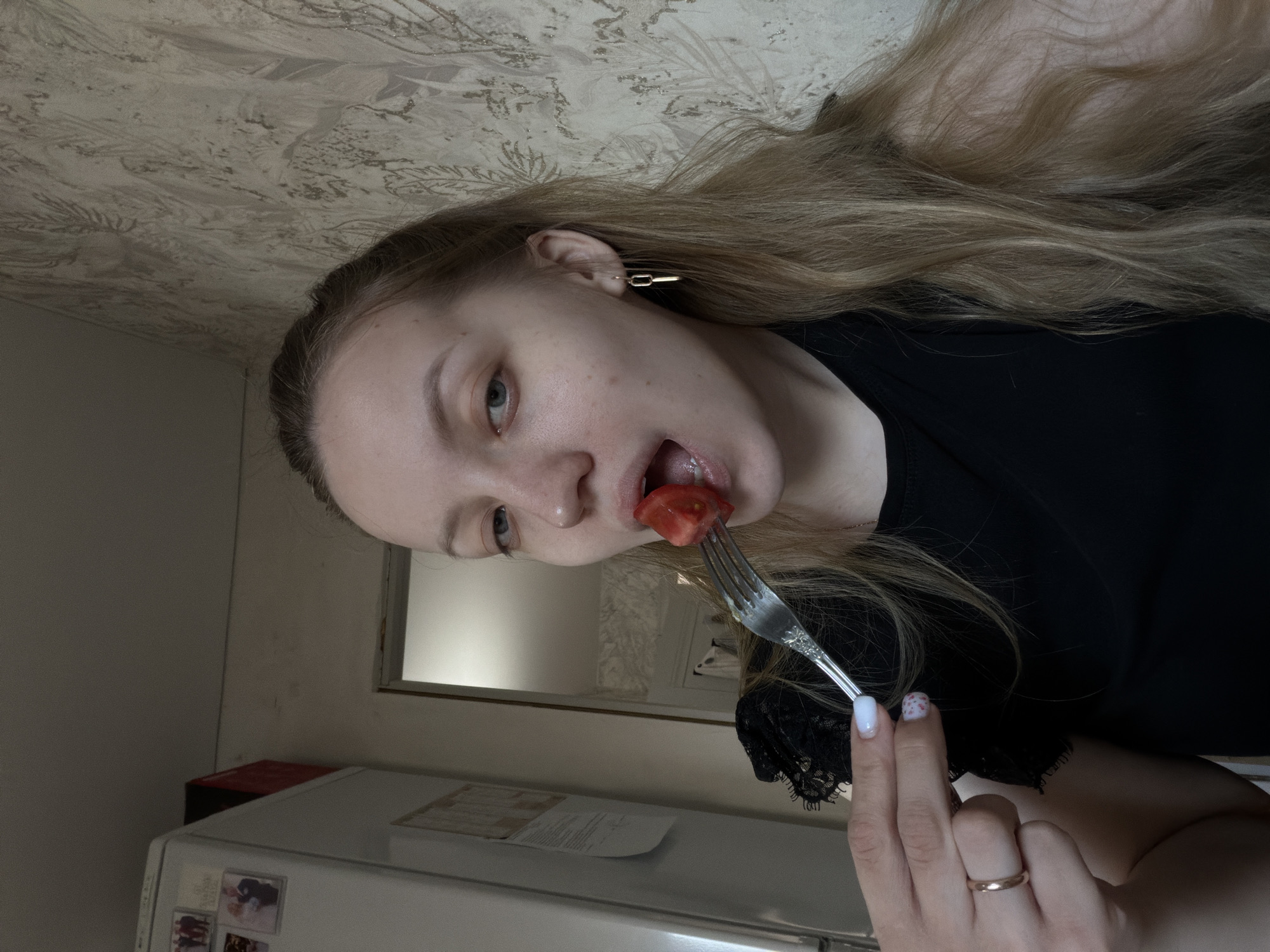 The scene depicts a young adult female indoors, in what appears to be a residential kitchen or dining area in Reshetnikovo, Russia. The female, possessing long blonde hair and light-colored eyes, is shown in the act of eating, holding a silver-colored fork with a red tomato wedge impaled on its tines directly in front of her open mouth. She is wearing a black top with a lace-detailed left shoulder, a gold-colored ring on the ring finger of her left hand, and a gold-colored rectangular link earring in her right ear. Her visible fingernails are manicured, with the thumb and index finger featuring light blue/white polish, and the middle finger displaying white polish with small red dots. In the background to the left, a white refrigerator is visible, adorned with multiple small framed photographs attached by magnets, including one depicting a wedding couple. A red container rests on top of the refrigerator. The wall behind and to the right of the subject is covered with light-toned, patterned wallpaper featuring feather or leaf motifs. A partially open doorway leading to another room is visible to the left of the subject's head. The ceiling is plain and light-colored.