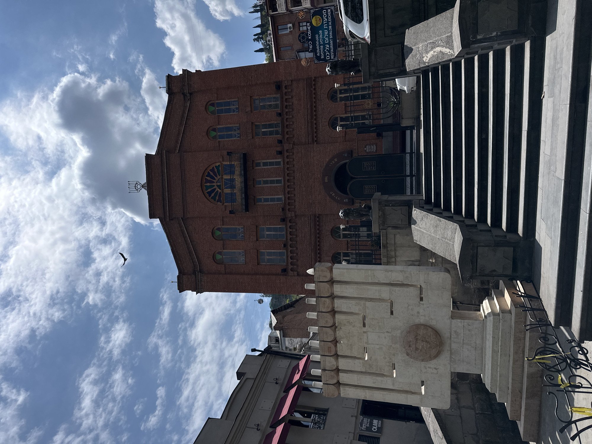 This media file captures an architectural scene, likely in Tbilisi, Georgia, on a bright, partly cloudy day. The central subject is a grand, multi-story building constructed of reddish-brown brick, which appears to be a synagogue. It features numerous windows, including several arched ones and a large, prominent round window above the main entrance, adorned with a blue and yellow stained-glass-like design. A metallic menorah sculpture is visible on the building's roof.
To the right of the synagogue, a wide, imposing stone staircase with heavy shadows ascends. In the lower left foreground, there is a substantial, light-colored stone monument or railing with a circular emblem carved into its surface and a stepped, castle-like top. Other buildings are visible in the background and to the left, including one with a red awning and signs.
Visible text on signs includes "כשר KOSHER" in both Hebrew and English, and "מסעדת שלום עליכם / RESTAURANT SHALOM ALEICHEM" in Hebrew, Georgian, and English, indicating a Jewish restaurant. Other signs read "OLAMI GEORGIA" and "ოლაში". Hebrew text, "שערי תפילה" (Gates of Prayer), is discernible above the synagogue's entrance. The sky is bright blue with scattered white and grey clouds, and a single bird is captured in flight. Yellow caution tape is tied to a railing in the lower right. The strong sunlight casts distinct shadows, suggesting daytime.