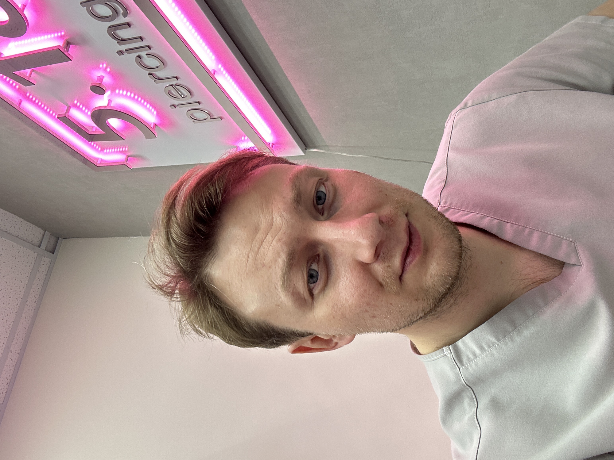 A fair-skinned man with short brown hair, blue eyes, and stubble is posing for a selfie. He is wearing a grey-blue scrub top. Above him is a sign with pink neon lettering that reads "5 Piercing". The location is likely Sochi, Russia, based on the sign's language. The environment appears to be a medical or body modification establishment.