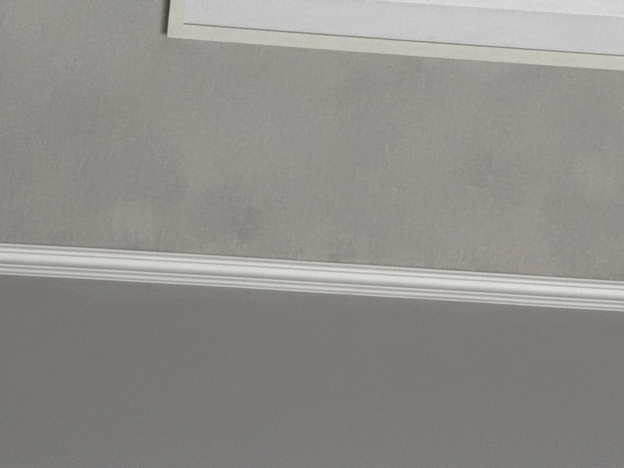 The image presents a close-up view of a wall, featuring a decorative molding along the lower part of the frame and a portion of what appears to be either a frame or the edge of a surface at the top. The wall is painted in a muted, neutral tone, exhibiting a subtle texture or pattern. The molding is white and has a classic design with horizontal lines. The lighting suggests that the scene is indoors, with the light source positioned to create shadows that subtly highlight the wall's texture and the molding's details. The overall composition is static and focuses on the architectural elements of the space. No people or identifiable objects are visible. The location is not discernible from the image itself.