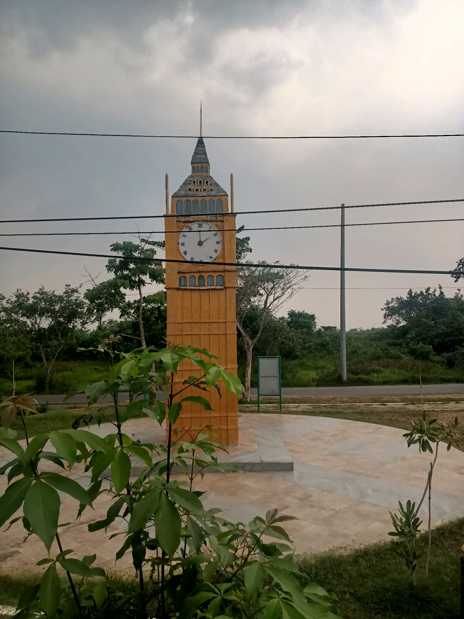 A tall, yellow, rectangular clock tower, resembling Big Ben, stands prominently in a public space. The tower features a clock face with Roman numerals and has decorative elements. The sky above is overcast with gray clouds. Several black wires stretch horizontally across the frame in the foreground and midground, obscuring parts of the tower and the sky. In the foreground, lush green foliage from plants partially obstructs the view of the tower and the paved ground. Behind the tower, a sparsely wooded area with a variety of trees and green foliage stretches towards the horizon. A paved pathway or road is visible behind the tower and to the left. A light pole stands to the right of the tower, with a green framed sign positioned near its base. The ground around the tower is paved with light-colored tiles arranged in a geometric pattern. This scene is located in the city of Bagus, Indonesia.