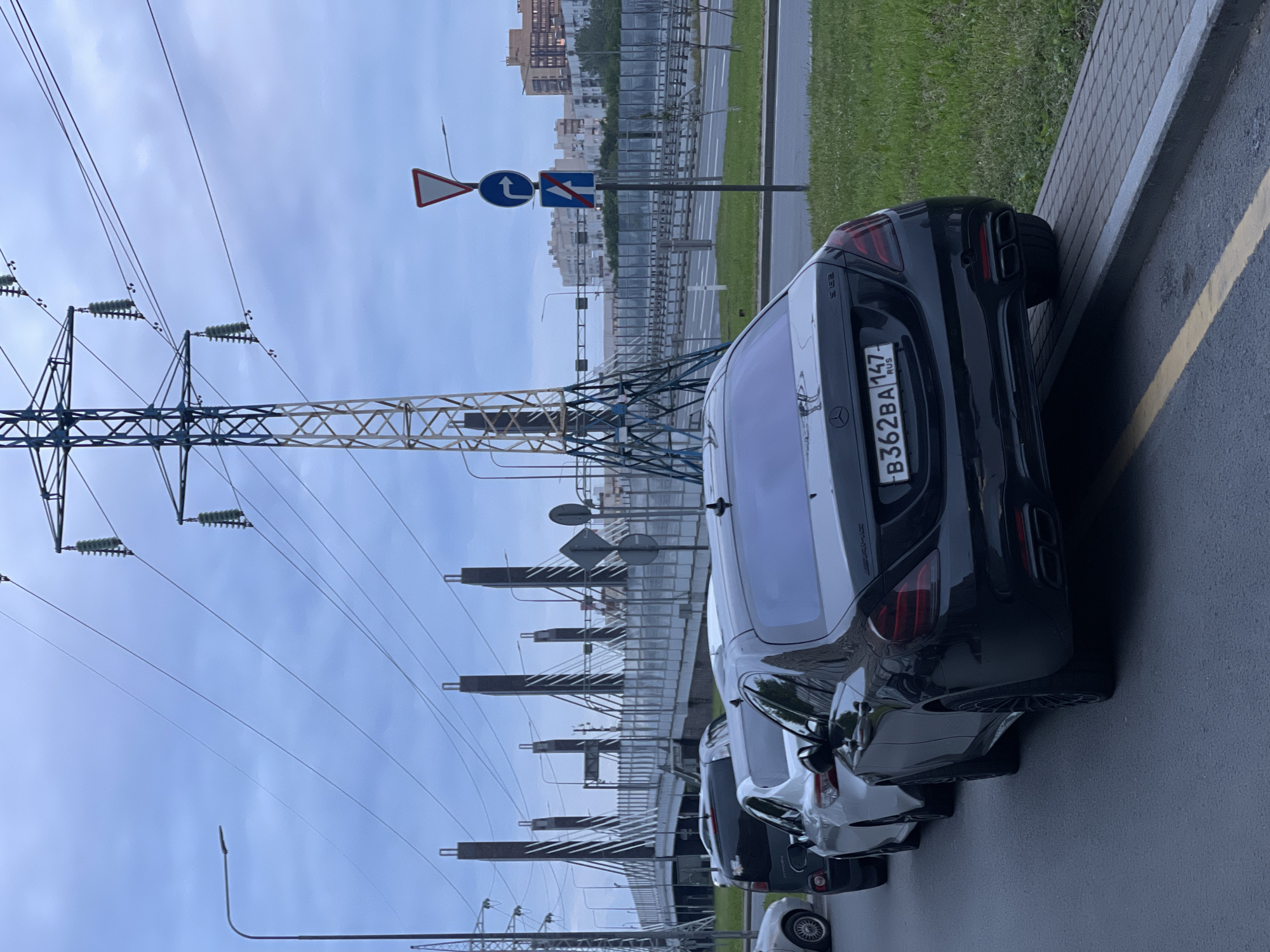 A black Mercedes-Benz S63 AMG sedan is parked on the side of a road, with its rear tires on a raised curb. The car has a Russian license plate that reads "B362BA 147". In front of the Mercedes are other parked cars, including a white sedan and a black SUV. In the background, a modern cable-stayed bridge with tall, dark-colored support towers spans across the scene. The sky is overcast, and there are large power lines and a tall metal transmission tower visible. Traffic signs, including a yield sign and a sign indicating a right turn and a straight lane, are posted on a pole to the right of the road. Residential buildings can be seen in the distance. The overall scene depicts a street view in a contemporary urban or suburban environment.