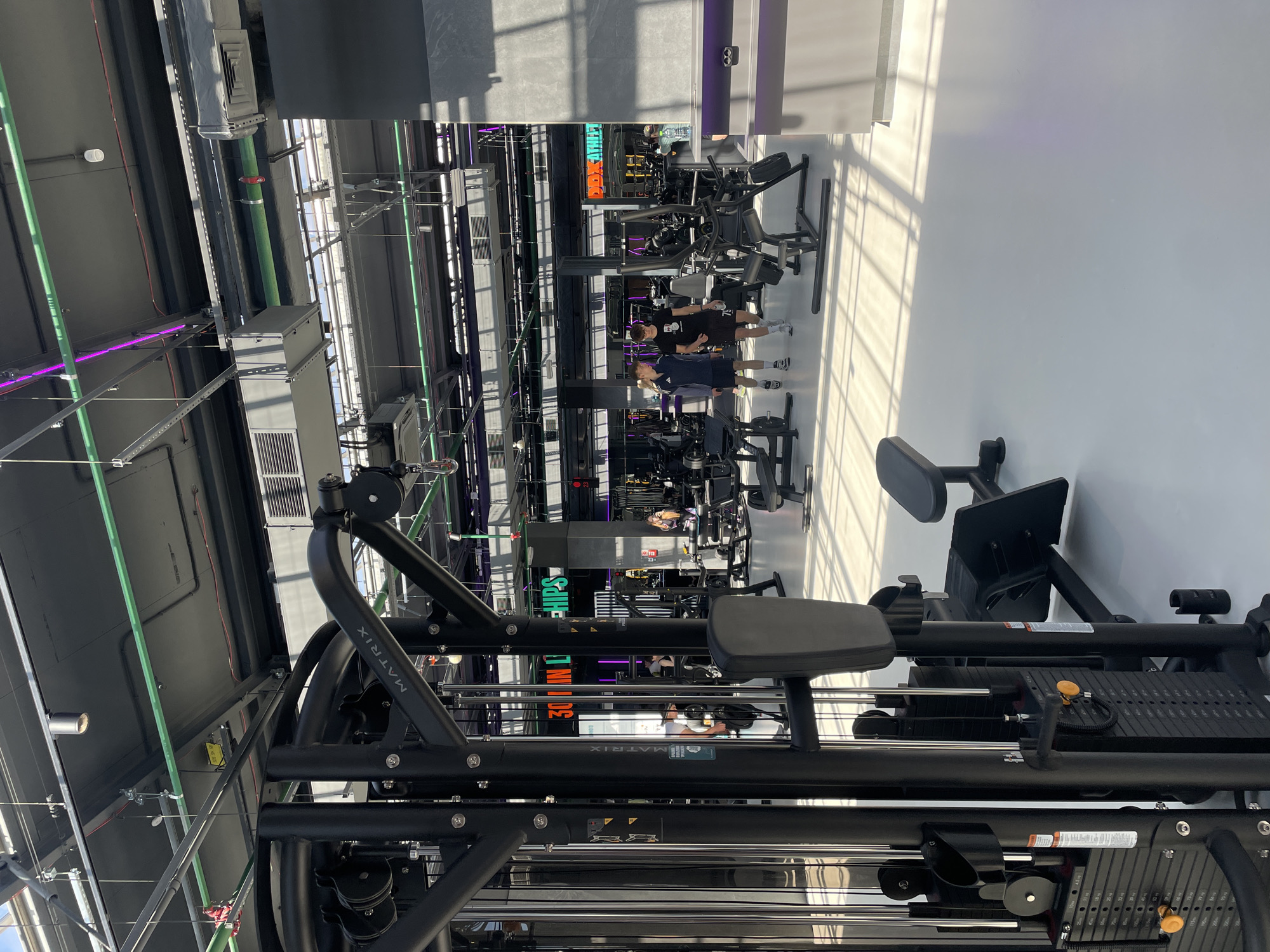 The image depicts the interior of a gym, featuring various workout machines and individuals engaged in physical exercise. The location is identified as Kazan, Russia.
Foreground: A black Matrix workout machine dominates the foreground, with a seat, weights, and various metal bars.
Midground: Several individuals are present in the midground, some appearing to be using other workout machines.
Background: The background reveals the gym's architecture, including a high ceiling with visible pipes and ventilation systems. Linear lighting fixtures, some emitting a purple hue, are also noticeable.
The overall environment appears to be well-lit and modern.