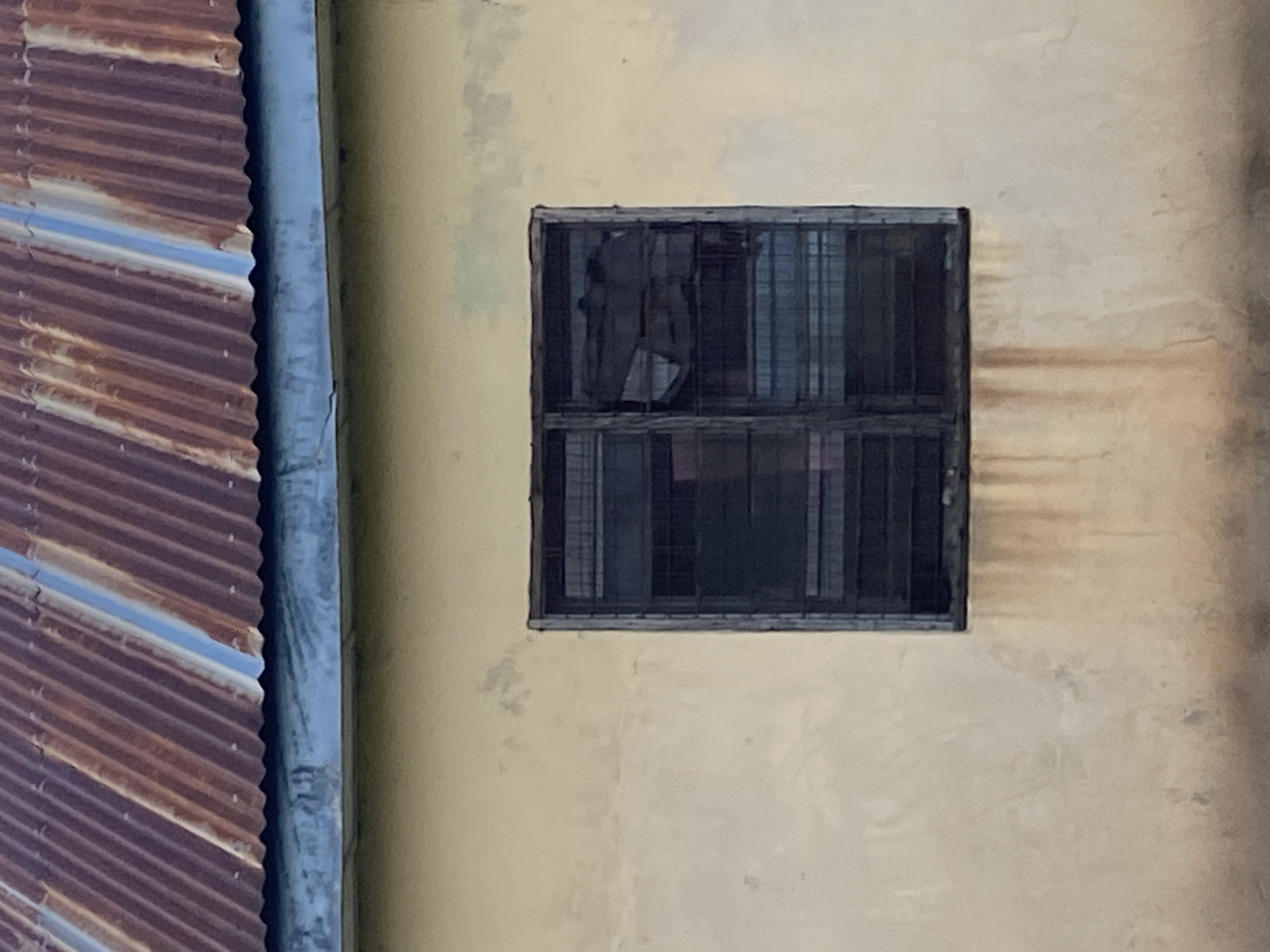 A medium shot captures the facade of a building under a corrugated metal roof in Jauro Yinu, Nigeria. The roof, showing signs of rust, has vertical ridges and some blue-tinted sections. Below the roofline, a light yellow wall forms the main body of the building. A dark, louvered window is centrally placed on the wall. The window is divided into two sections. The left section is dark, appearing opaque, while the right section has a broken pane, revealing darkness and some obscured shapes within. Water stains streak down the yellow wall beneath the window. The environment appears to be outdoors, with hints of shadows and muted colors suggesting it could be daytime. There are no people visible in the image.