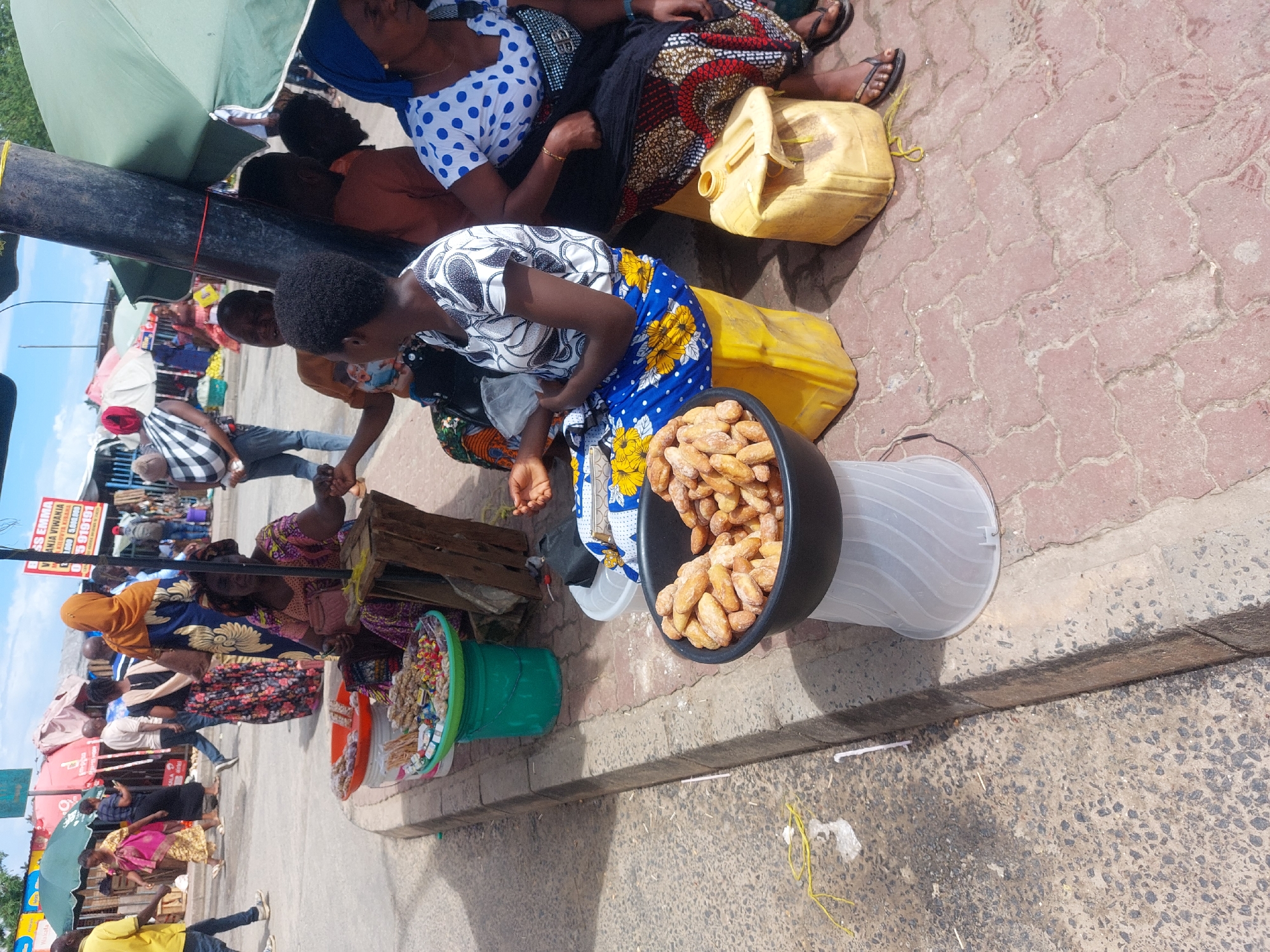 The image depicts a bustling street market scene in Dar es-Salaam, Tanzania, under bright daylight. The setting is a paved sidewalk, made of red interlocking bricks, running alongside a street.
In the foreground, a woman wearing a black and white patterned top and a blue skirt with yellow floral designs is seated on a yellow jerrycan. She appears to be a vendor, with a large black bowl in front of her filled with golden-brown fried dough snacks. To her left, a green bucket holds a variety of colorful packaged candies and snacks. Another woman, dressed in a white polka-dot top and a patterned skirt, sits further left under a large green market umbrella, also appearing to be a vendor. Several yellow jerrycans are scattered around, used as seats or storage.
Beyond these foreground figures, the street is active with numerous people walking and standing, suggesting a lively market environment. Other market stalls and structures are visible along the street, with additional umbrellas providing shade. A black pole stands upright near the main green umbrella, likely part of the market infrastructure.
The bright sunlight indicates it is daytime, possibly midday or early afternoon, with clear shadows cast. The overall atmosphere is one of busy commerce. On a partially visible sign in the background, the text "MISS EMMA" and "AKIA VIWANJA" along with the numbers "919191" can be seen, likely advertising a local business.