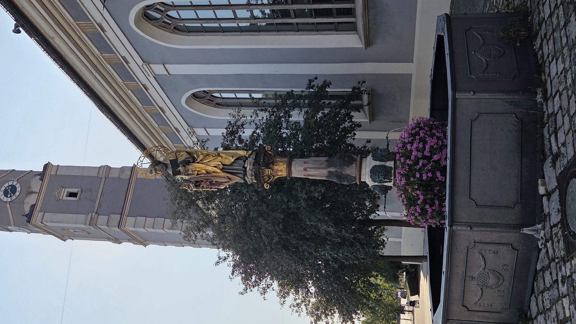 A golden statue of a saint, holding a book and a halo, stands atop a column in a fountain in Ulm, Germany. The statue is positioned in front of a building with a gray facade and arched windows, and to the left of a clock tower. The fountain basin is octagonal and made of dark metal, decorated with relief patterns. Pink flowers bloom within the fountain, surrounding the base of the statue. The ground is paved with cobblestones. In the background, trees line a street where a person in a wheelchair is visible. The sky is clear and bright.