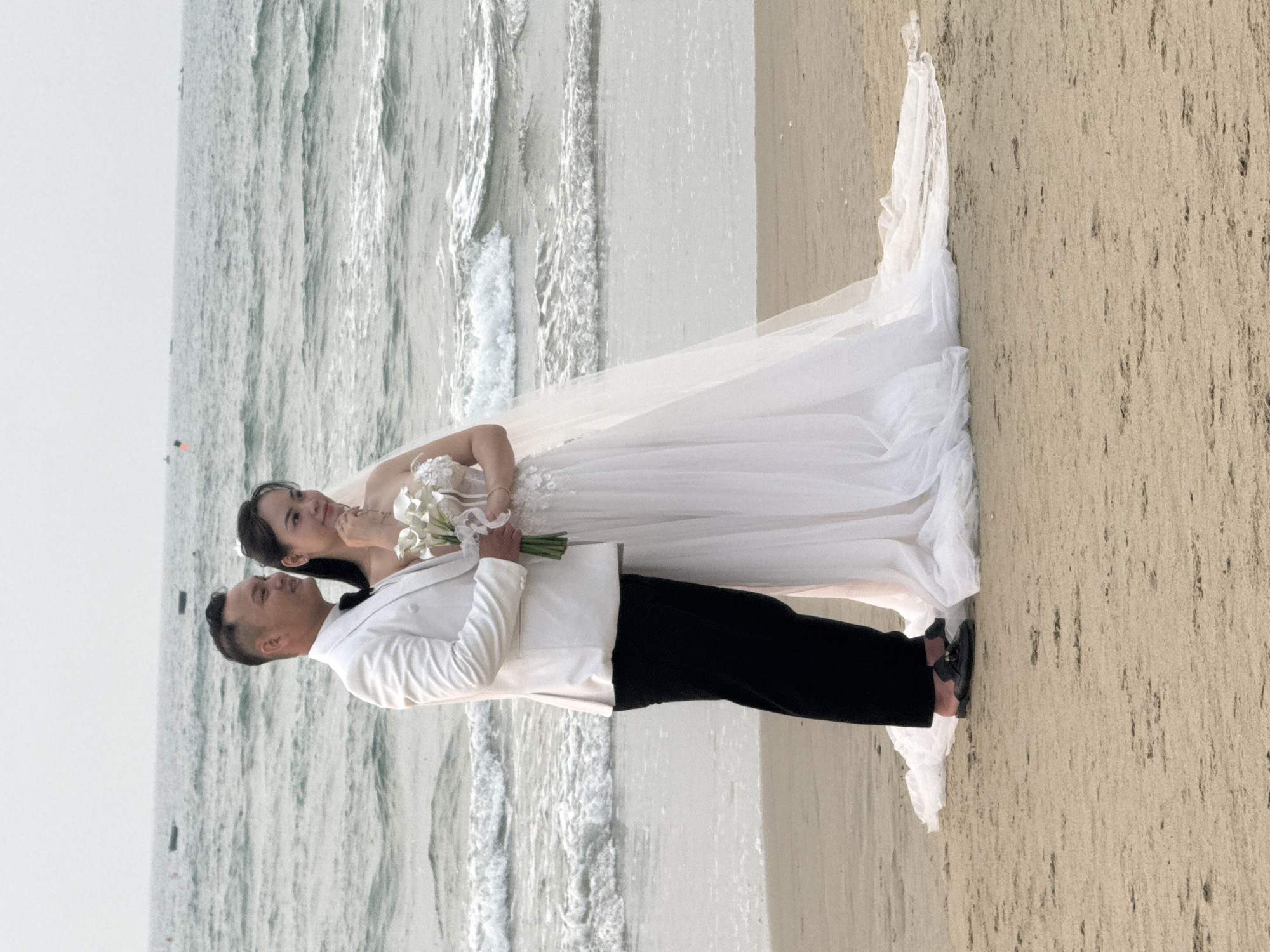 A wedding photo of a couple on a beach in Đà Nẵng, Vietnam. The bride is wearing a white wedding dress and holding a bouquet of white lilies, while the groom is wearing a white jacket and black pants. They stand together facing the ocean, which is visible in the background, along with a sandy beach. The sky is overcast.