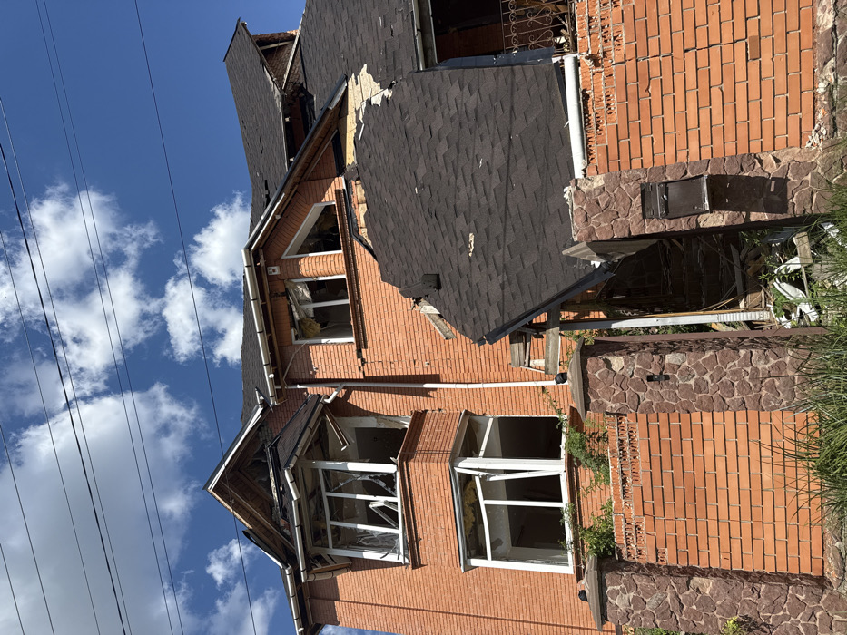 The image depicts a two-story residential building in Markhalivka, Ukraine, exhibiting significant damage under a clear blue sky with scattered white clouds. The structure is constructed of red brick, with the primary damage concentrated on the roof and windows.
The dark-shingled roof shows extensive impact, with large sections torn, displaced, or completely missing, exposing underlying wooden structures and insulation. A prominent portion of the roof on the right side of the image appears to have collapsed outwards and downwards from the main structure. Multiple windows on both the upper and lower floors are shattered, with glass absent or fragmented, and frames distorted or dislodged. Visible through these openings are internal debris and insulation material. A bay window structure on the upper floor is heavily damaged, its own mini-roof section partially collapsed. White guttering and downspouts are visible along the eaves, some appearing intact while others may be compromised.
In the foreground, a low perimeter fence constructed of red brick with dark stone-clad pillars runs horizontally. A segment of this fence near the center is visibly damaged or collapsed, revealing rubble and debris behind it, including what appears to be discarded household items or structural wreckage. A dark-colored mailbox is affixed to one of the stone pillars. Green vegetation is present behind the fence and around the base of the house. Overhead, several dark utility lines traverse the upper portion of the frame against the sky. No individuals or animals are present within the scene.