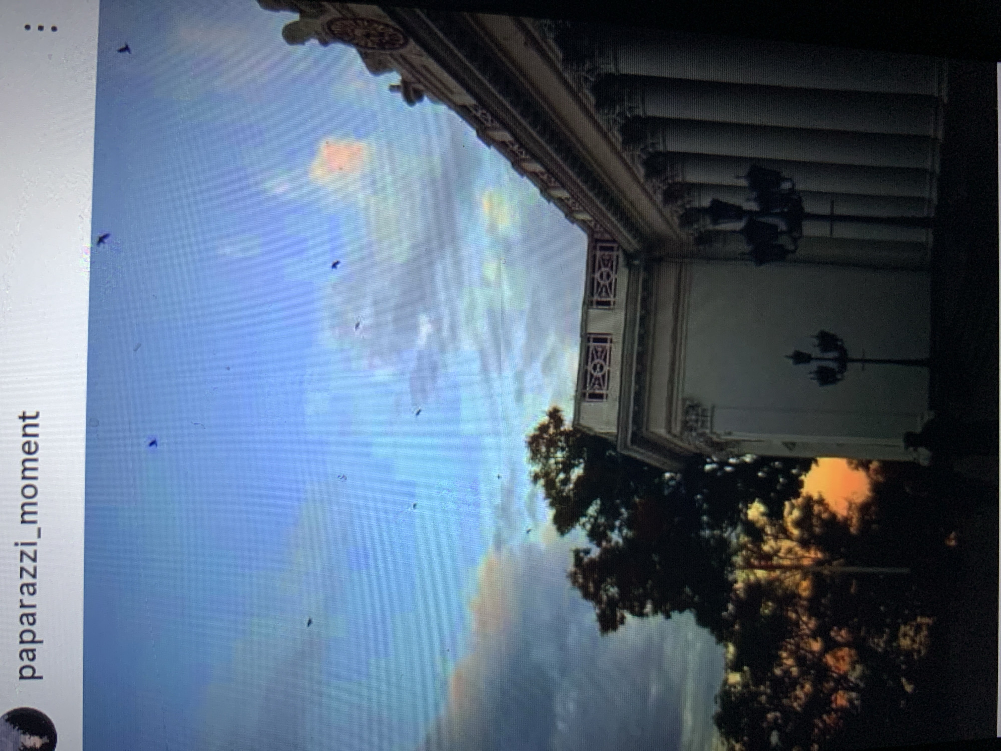 The image shows a building with classical architecture, featuring columns and ornate detailing. A large tree is visible to the left of the building. The sky is a mix of blue and grey clouds, with hints of orange from a sunset. Several small, dark shapes that appear to be birds are scattered across the sky. The text "paparazzi_moment" is visible at the top of the image, suggesting it is from a social media post. The location is identified as Lymanka, Ukraine.