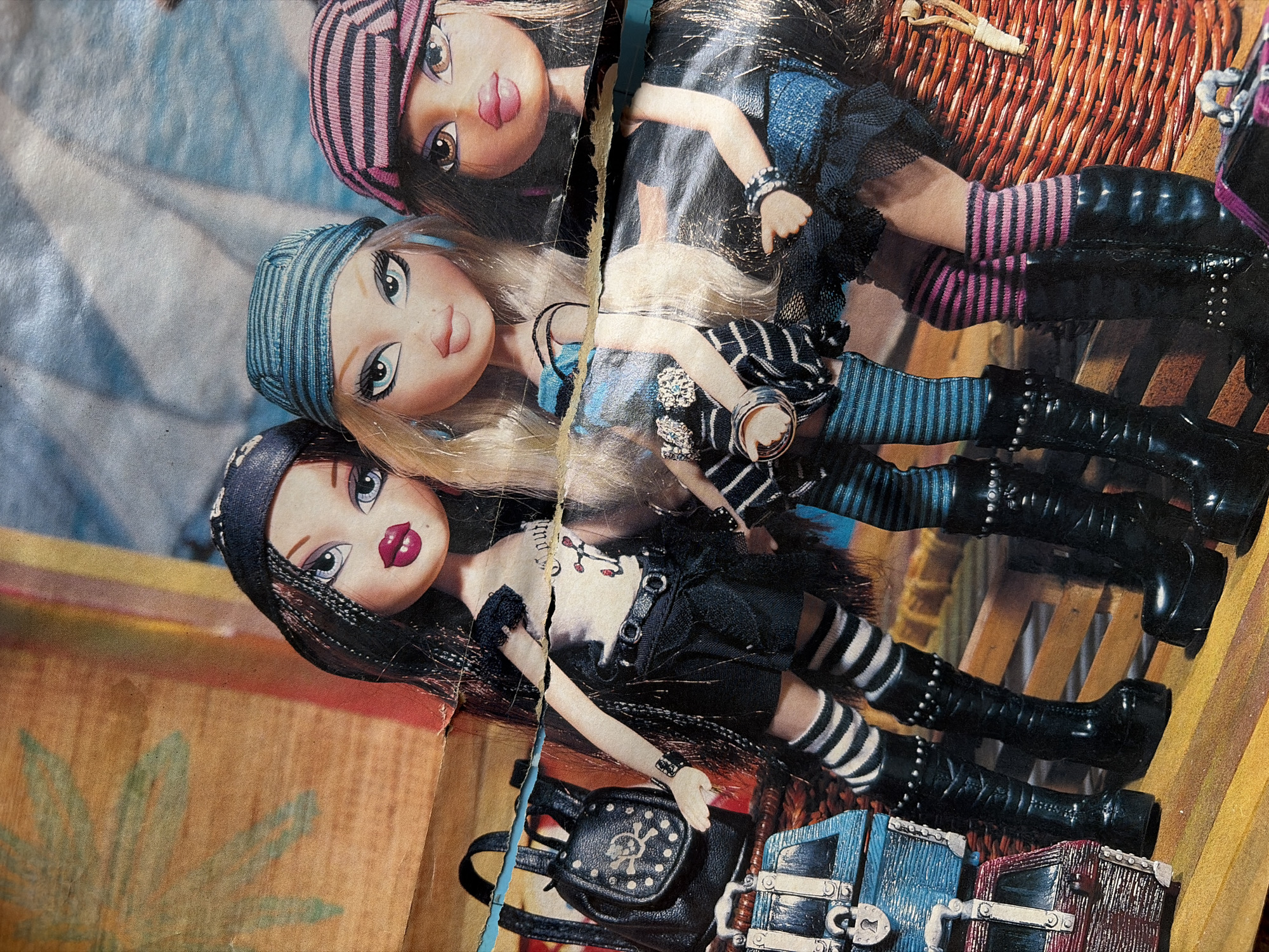 Three Bratz dolls are posed in a static arrangement, appearing to be part of a display or advertisement. The dolls are dressed in pirate-themed attire, including bandanas, striped socks, and tall black boots. One doll on the left has long black hair and wears a black skirt and t-shirt with a skull and crossbones graphic. The middle doll has blonde hair and wears a blue and black striped outfit. The doll on the right has brown hair and is dressed in a blue top and skirt with a pink and black striped bandana. Accessories such as a black backpack with a skull detail and two decorative chests are visible in the foreground. The background appears to be a collage of textures and colors, possibly depicting a beach or nautical theme, with elements like a palm frond and sail-like shapes. The overall image has a worn, slightly crumpled appearance, suggesting it might be a printed item like a poster or magazine page. The location of Malyshevo, Russia is not depicted or implied in the image.