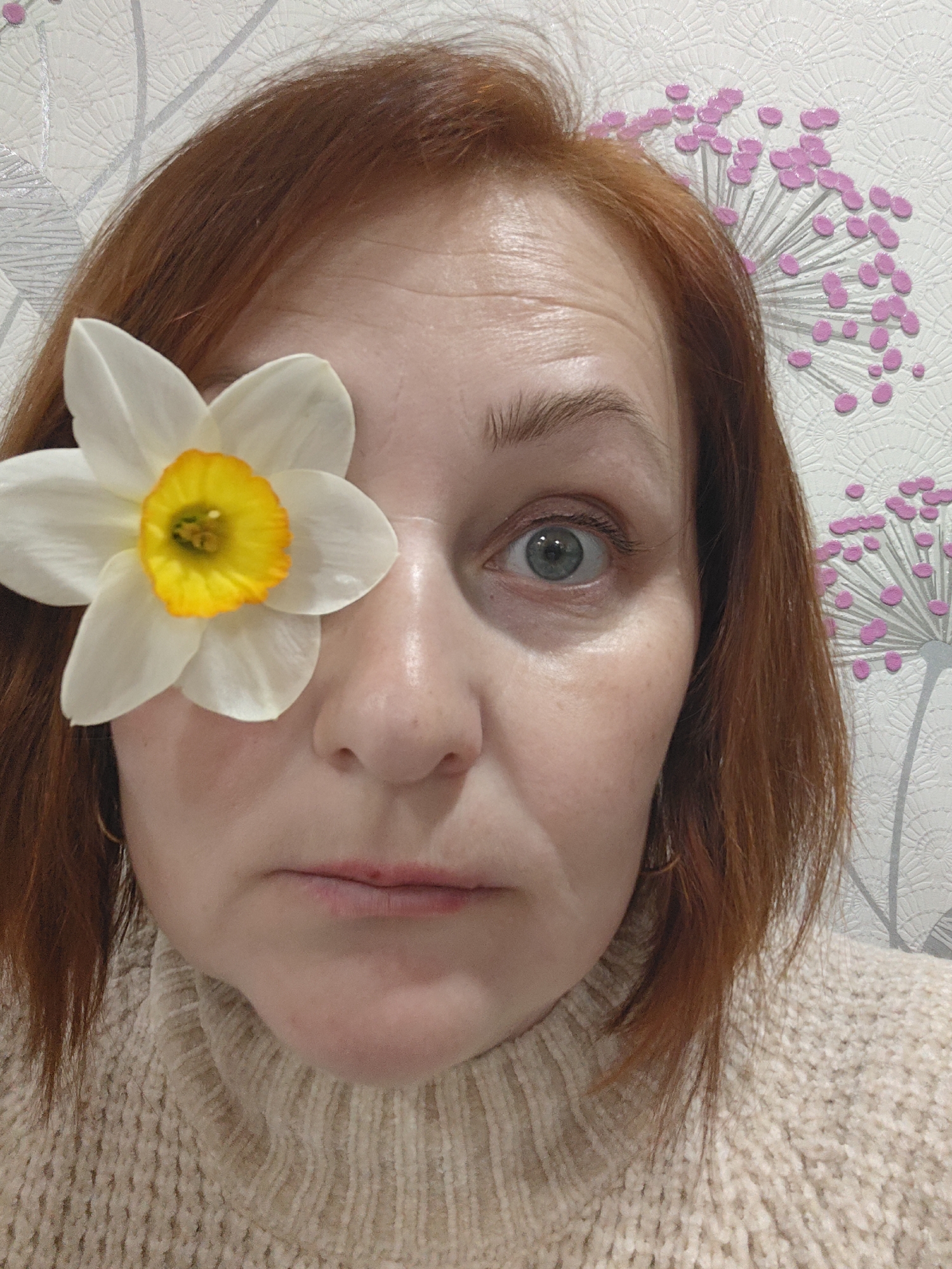 A middle-aged woman with red hair is posing for a selfie. A daffodil flower covers her left eye. She wears a cream-colored turtleneck sweater. The background is a white wallpaper with pink and gray floral patterns. The image was taken indoors. The location is not deducible from the given information.