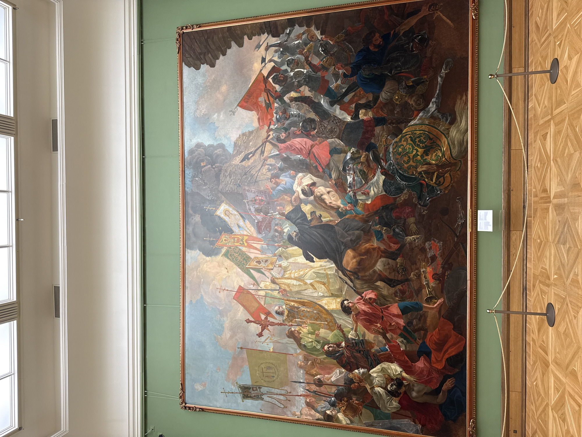 A large, vertically oriented painting depicting a historical battle scene hangs on a green wall inside a building. The painting is framed with an ornate gold frame. The battle scene features numerous figures engaged in combat, including soldiers, horses, flags, and weaponry. The colors are rich and varied, with prominent reds, blues, and golds. The composition is dynamic, with a sense of movement and chaos. The painting is lit by natural light coming from a nearby window visible on the left. A museum barrier is on the right, held up by a metal rod. The pattern of wood on the right appears to be herringbone or diamond shapes. The location is likely a museum or historical site within the city of Moscow, Russia.