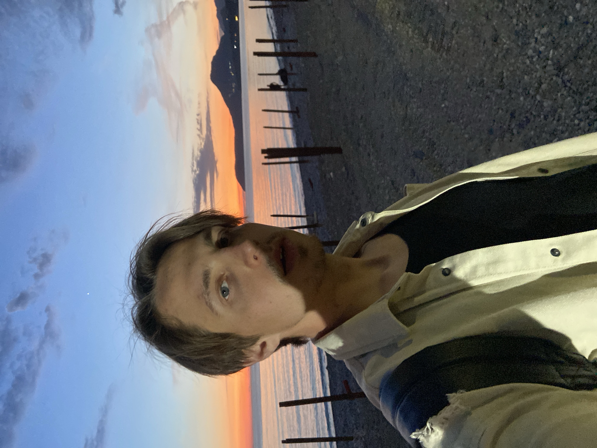 A young man captures a selfie on a beach at sunset in Bar, Montenegro. The subject, positioned on the right side of the frame, looks directly at the camera with blue eyes and a neutral expression, wearing a light-colored collared jacket over a dark t-shirt. A hint of stubble is visible on his chin.
The background reveals a picturesque coastal scene during twilight. The sky, dominating the left portion of the image, transitions from soft blue with scattered clouds at the top to vibrant orange and pink hues near the horizon, indicative of a sunset or sunrise. A dark silhouette of distant mountains or hills stretches across the horizon line. Below the mountains, the calm body of water, likely the Adriatic Sea, reflects the vivid colors of the sky in elongated streaks of orange and pink.
The immediate foreground and middle ground feature a dark, pebbly or gravelly beach. Several dark, weathered wooden posts or pylons emerge from the water near the shore, adding a structural element to the natural landscape. Faint lights can be discerned on the distant mountain slopes, suggesting the presence of buildings or settlements. The overall mood is peaceful and serene, with no discernible text in the image.