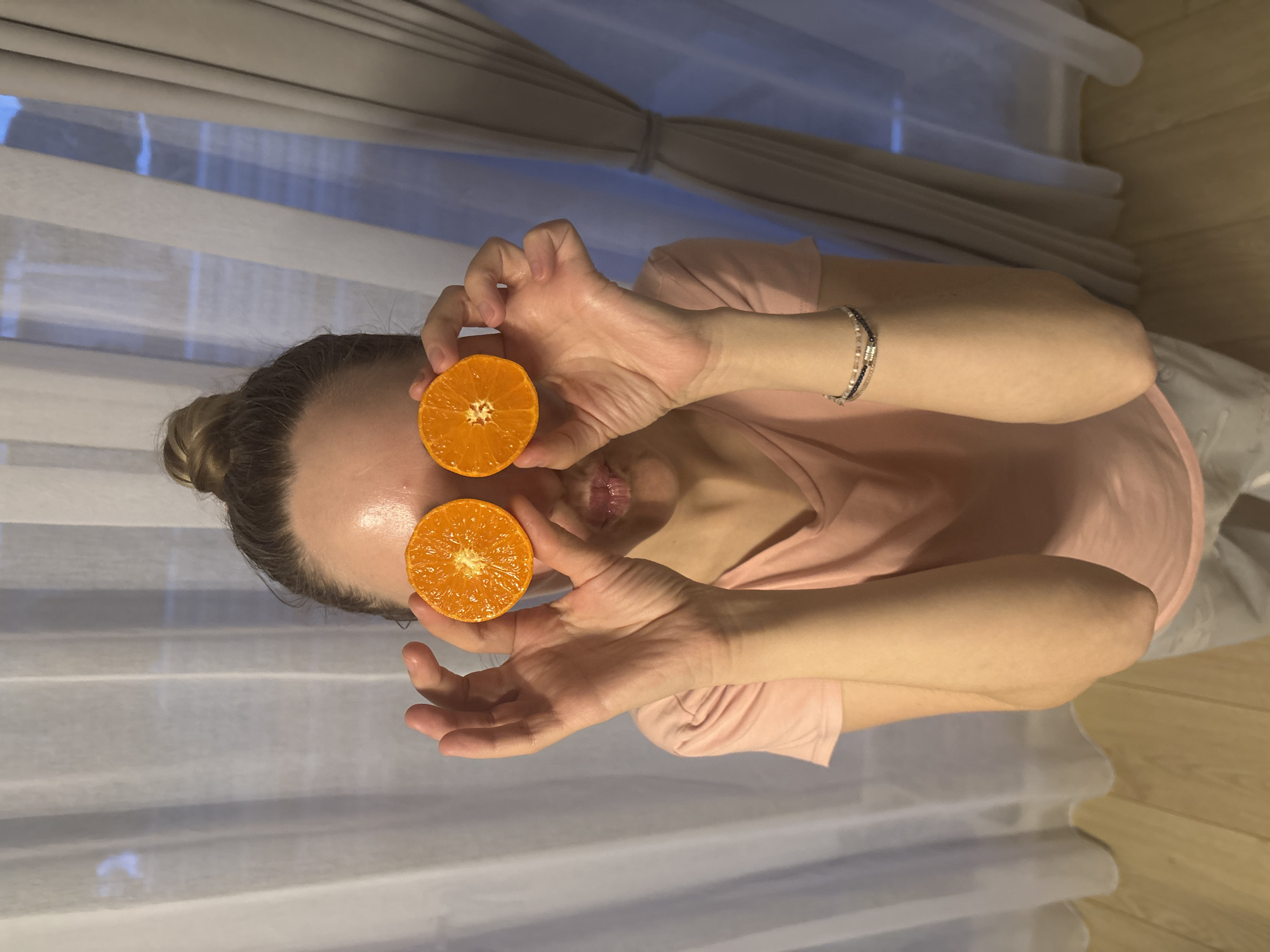 A woman is holding two orange halves over her eyes indoors. She is wearing a pink t-shirt and a bracelet. The background consists of a window with sheer white curtains and a wooden floor. The location is Chaikovsky, Russia.