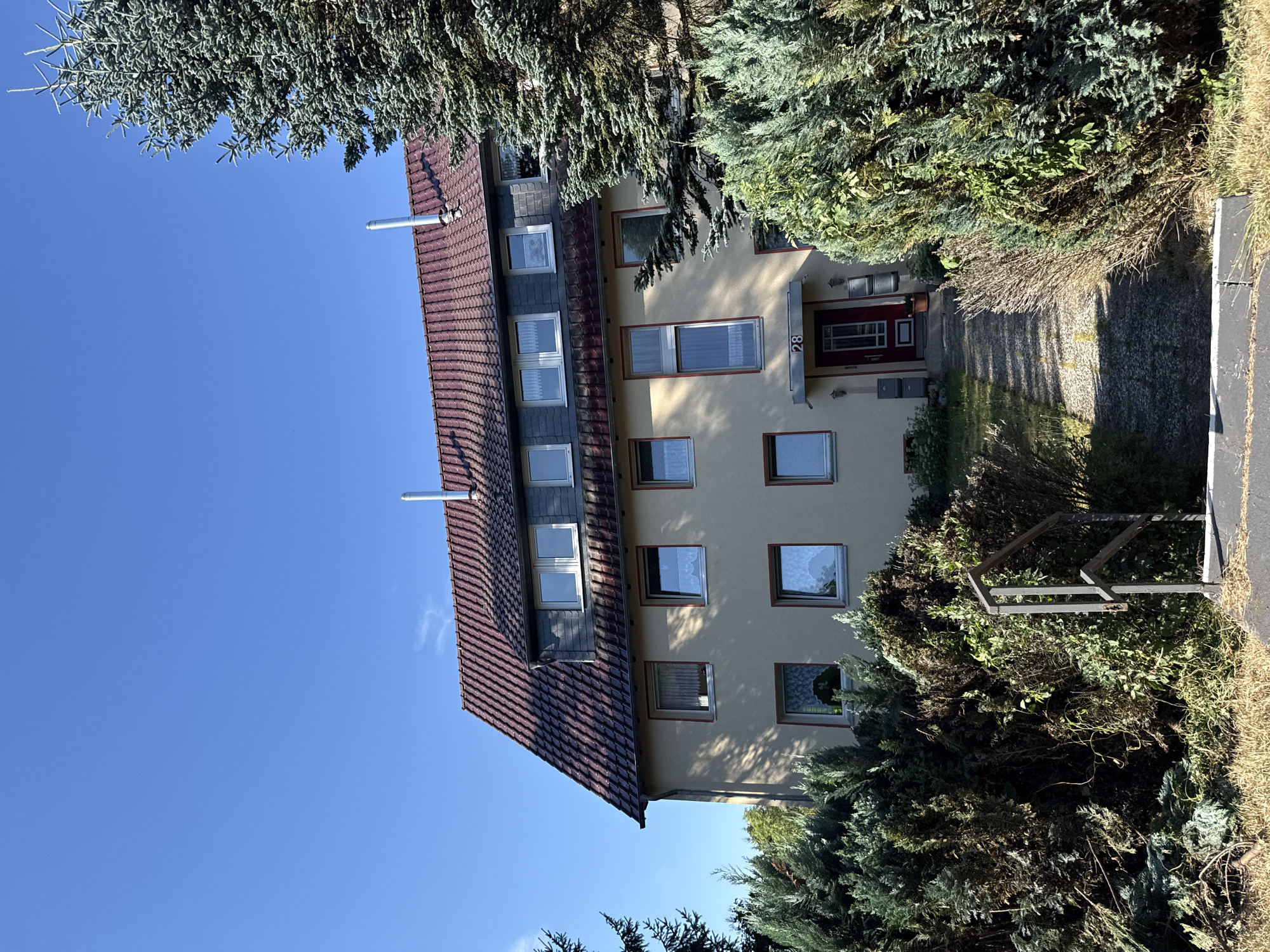 The image depicts the exterior of a multi-story residential building with a red tiled roof under a clear blue sky. The building has a light-colored facade and numerous windows. In front of the building, there is a path leading to a red door with the number "28" above it. Several mailboxes are mounted on the wall next to the door. Dense evergreen bushes and trees surround the building, with some areas appearing overgrown. The foreground shows a gravel path and a metal railing. The scene appears to be in Walkenried, Germany. There are no people visible in the image.