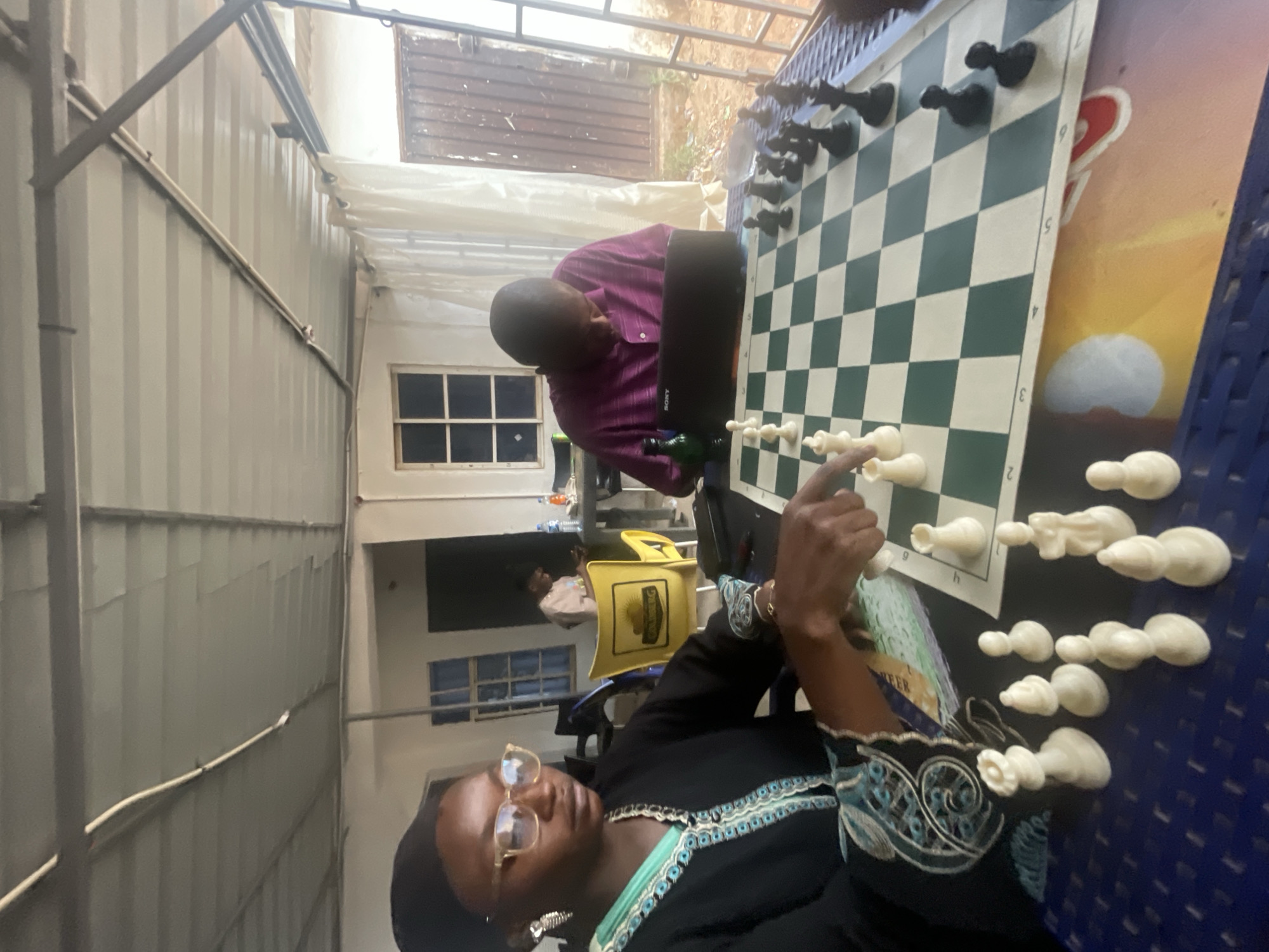 Two people are playing chess outdoors in what appears to be a covered courtyard. A person in a black shirt with blue trim is reaching toward the chessboard with their right hand. Another person in a purple shirt is sitting opposite them. The chessboard is set up on a blue patterned table. The surroundings include a corrugated metal wall, a window, and various chairs, including a yellow "Gulderberg" branded chair. A window and a gate are partially visible in the background. The location is Barakin Akawo, Nigeria.