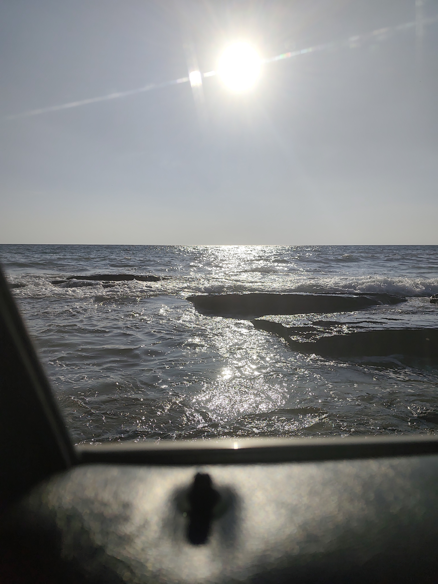 This media file is a photograph of a seascape with the sun shining brightly in the sky. The scene depicts a body of water, likely the sea, with waves gently rolling onto a rocky shore. The sun, positioned high in the sky, creates a brilliant glare and reflects off the water's surface, casting shimmering highlights. The sky is clear and pale blue, with a thin, elongated cloud streak visible to the left of the sun. In the foreground, out of focus, is a dark, metallic object with a visible bolt or screw, suggesting the photograph may have been taken from a boat, a pier, or some other structure overlooking the water. The lighting suggests it is daytime, likely late morning or early afternoon, given the sun's high position and the intensity of its rays. The overall mood is serene and peaceful, with the natural beauty of the ocean and the bright sunlight creating a calming atmosphere. There is no visible text in the image.