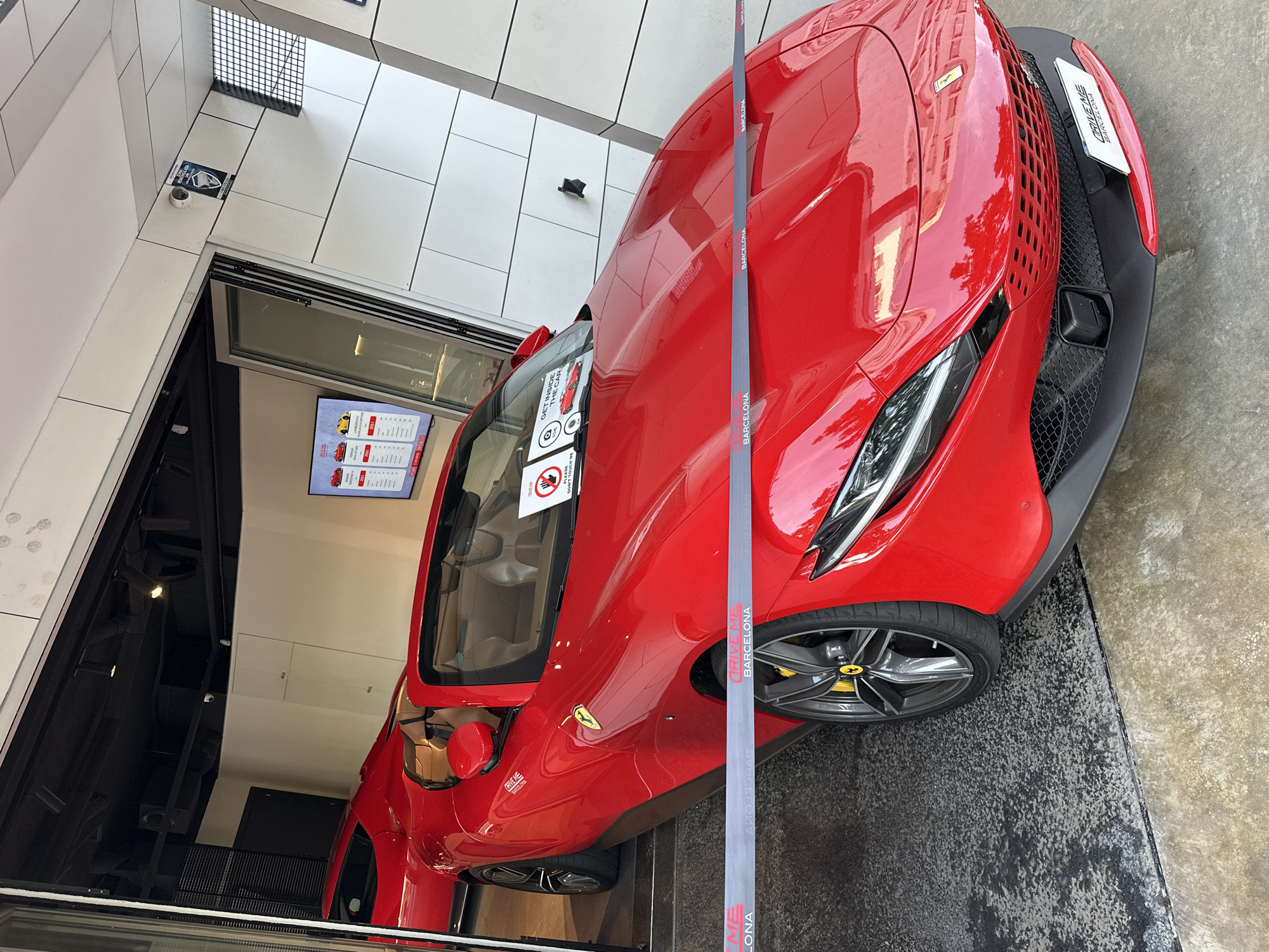 A red Ferrari Roma Spider is prominently displayed at what appears to be a luxury car rental or experience showroom in Barcelona, Spain. The vehicle, a convertible with a light tan interior, is positioned so its front half is outdoors on a speckled dark asphalt surface, while its rear half is elevated on a step inside the showroom, which features white paneled walls and a black ceiling with track lighting.
A grey ribbon or barrier, emblazoned with the repeated text "DRIVE ME BARCELONA" in red and white lettering, stretches horizontally across the front quarter panel and hood of the Ferrari, preventing close access. On the windshield, two white circular signs are visible: one with a red "no touching" symbol and "PLEASE DON'T TOUCH ME" text, and another with a camera icon and the text "GET INSIDE THE CAR." The Ferrari's wheels are dark grey with yellow brake calipers, and the iconic yellow Ferrari emblem is visible on the front quarter panel and on the center caps of the wheels.
Inside the showroom, partially visible behind the lead Ferrari, is another red sports car, also presumably a Ferrari. A large digital screen on the back wall displays information about various car models available for driving experiences, including a red Ferrari and a yellow Lamborghini, with pricing in euros. The exterior of the building features white geometric panels and a large glass folding door system. A security camera is mounted on the white facade above the entrance, and a blue and white sign, possibly indicating security or COVID-19 protocols, is affixed near it. The overall scene suggests a retail or experiential setting for high-end automobiles.
