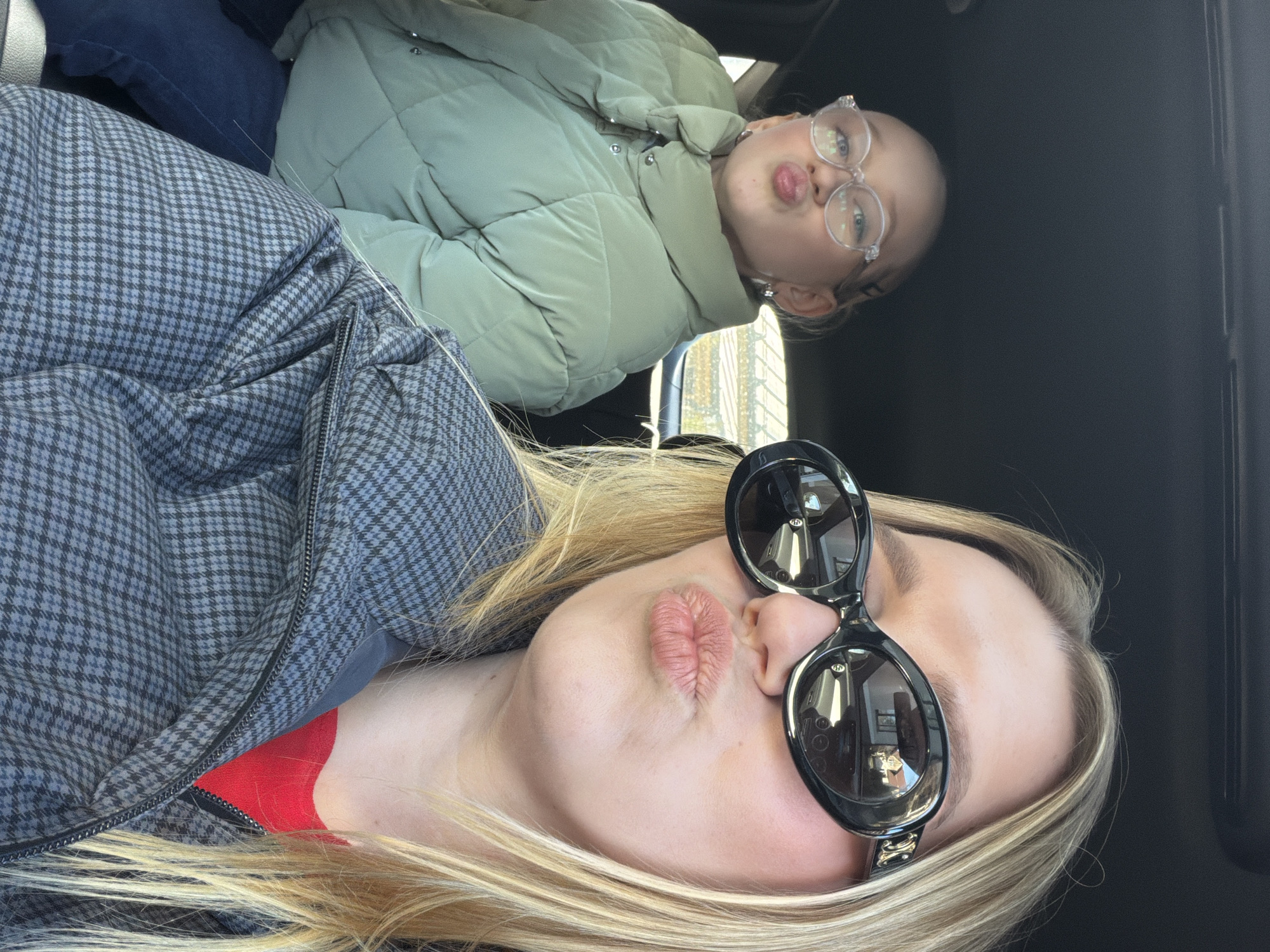 Two fair-skinned individuals are in a vehicle. One is wearing sunglasses and a houndstooth jacket, with a red garment visible underneath. The other person wears eyeglasses and a light green puffer jacket. Both are making a kissing face. The vehicle's interior is mostly black. The location is plausibly Dreiliņi, Latvia, based on the visible outdoor scenery through the car window, suggesting an urban residential area.